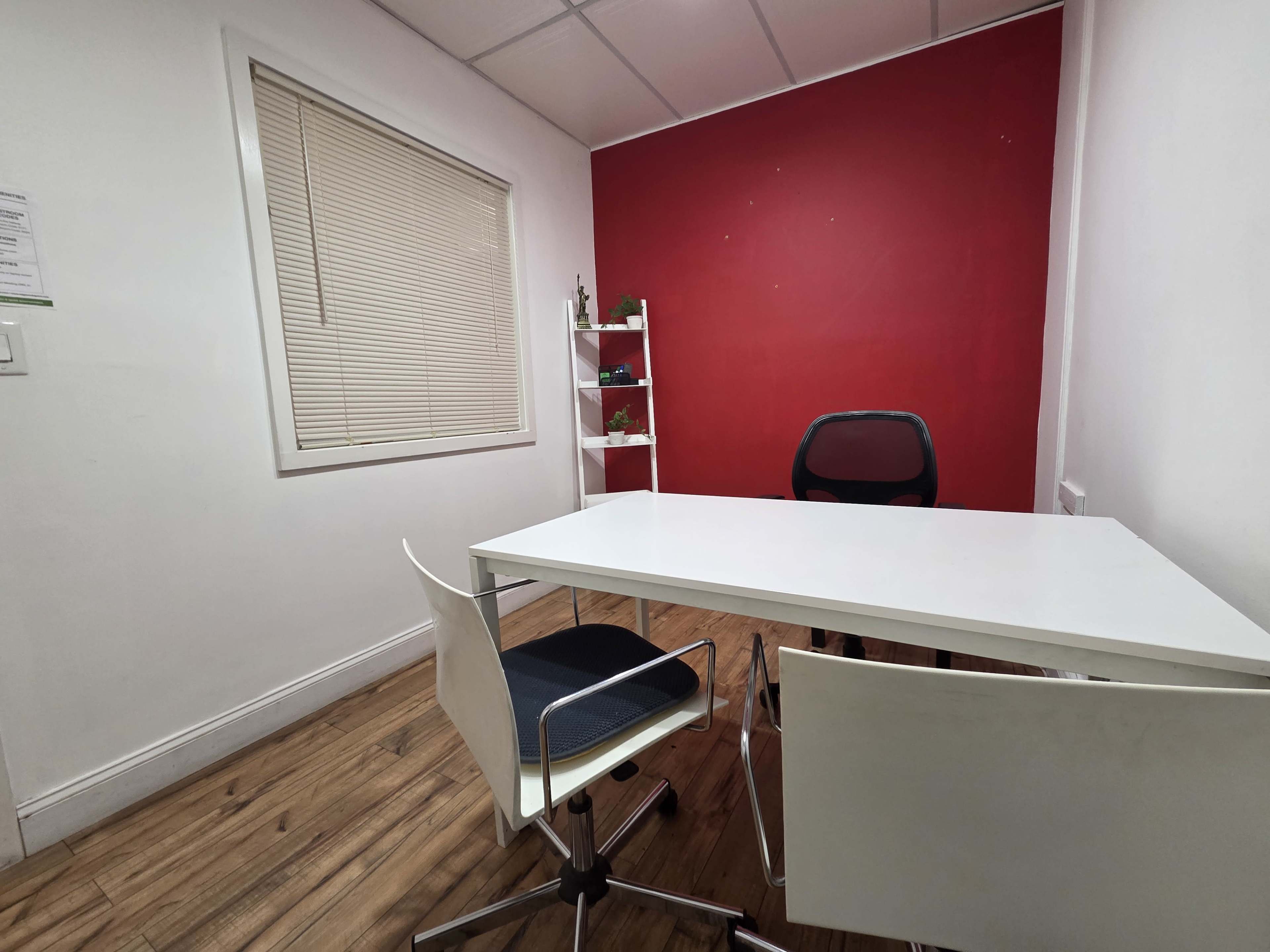 The image shows a small office room with a white table, two chairs, and a red accent wall, along with a shelving unit.