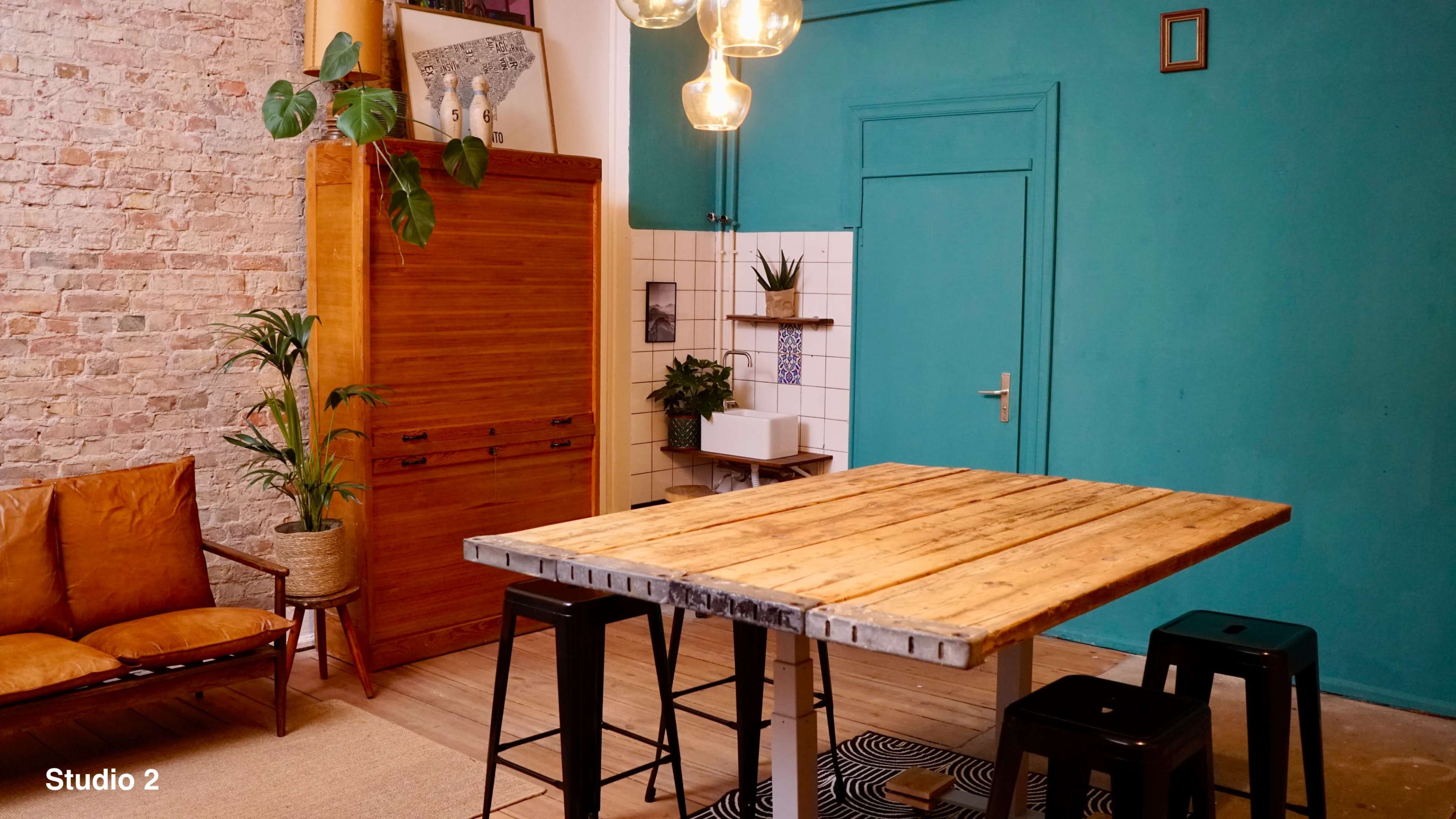 The image shows a room with a wooden table surrounded by black stools, featuring a turquoise wall, plants, and various decorative elements.