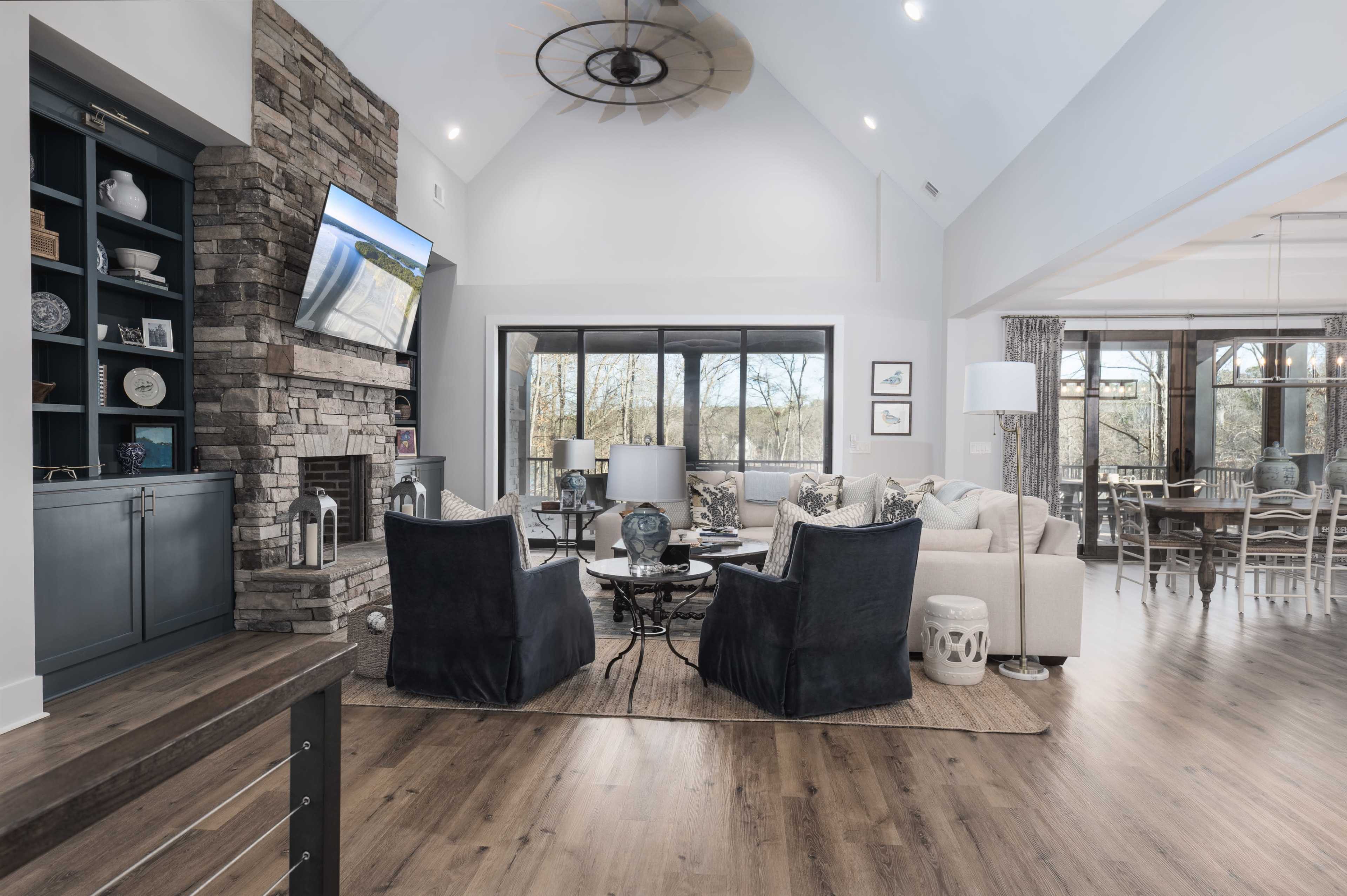 The image shows a modern, airy living room with a stone fireplace, a large television mounted on the wall, and a mix of comfortable seating arrangements including two armchairs and a sofa.
