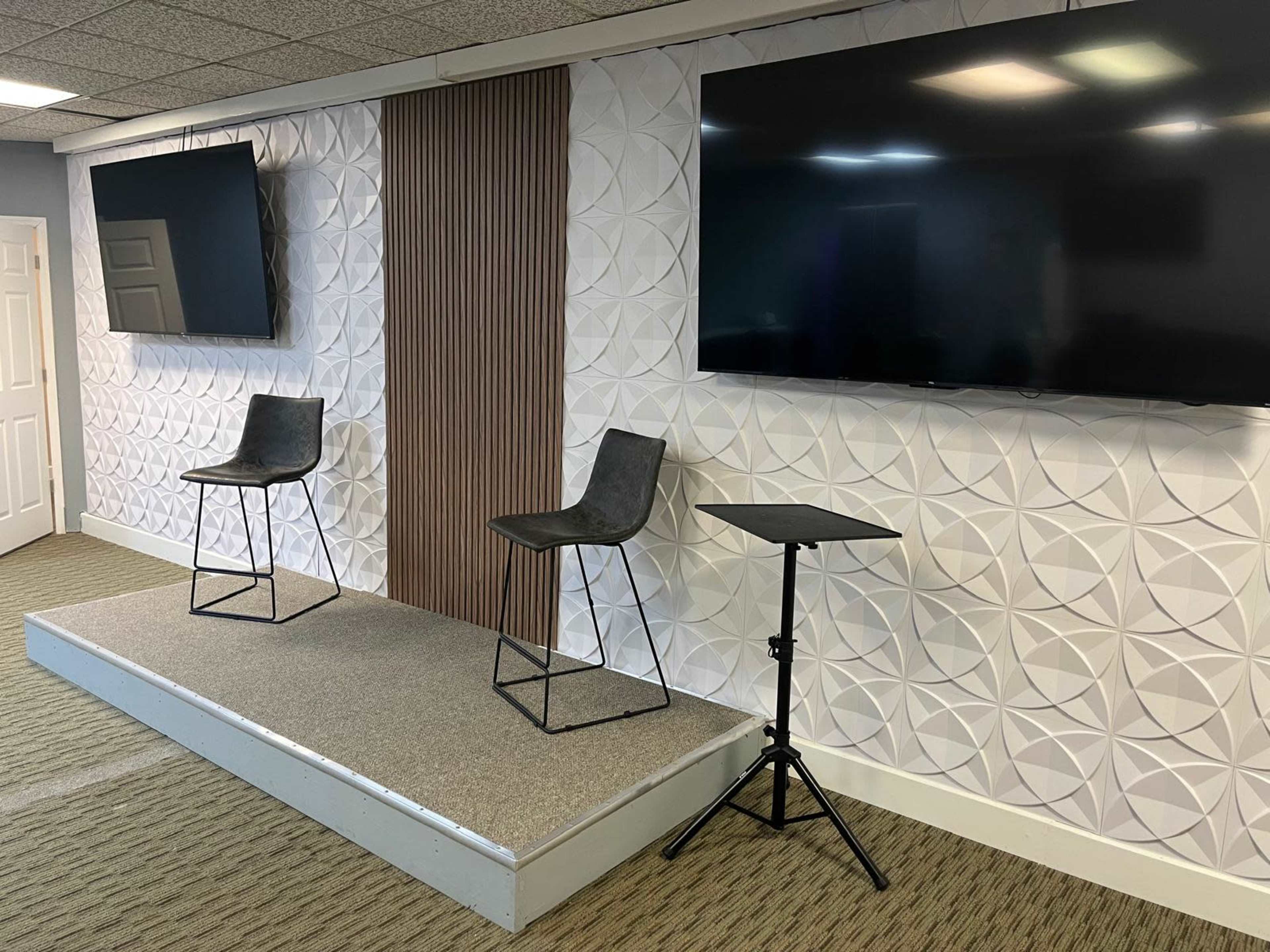 The image shows a presentation stage with two black chairs and a small table, set against a textured wall featuring two large screens on either side.