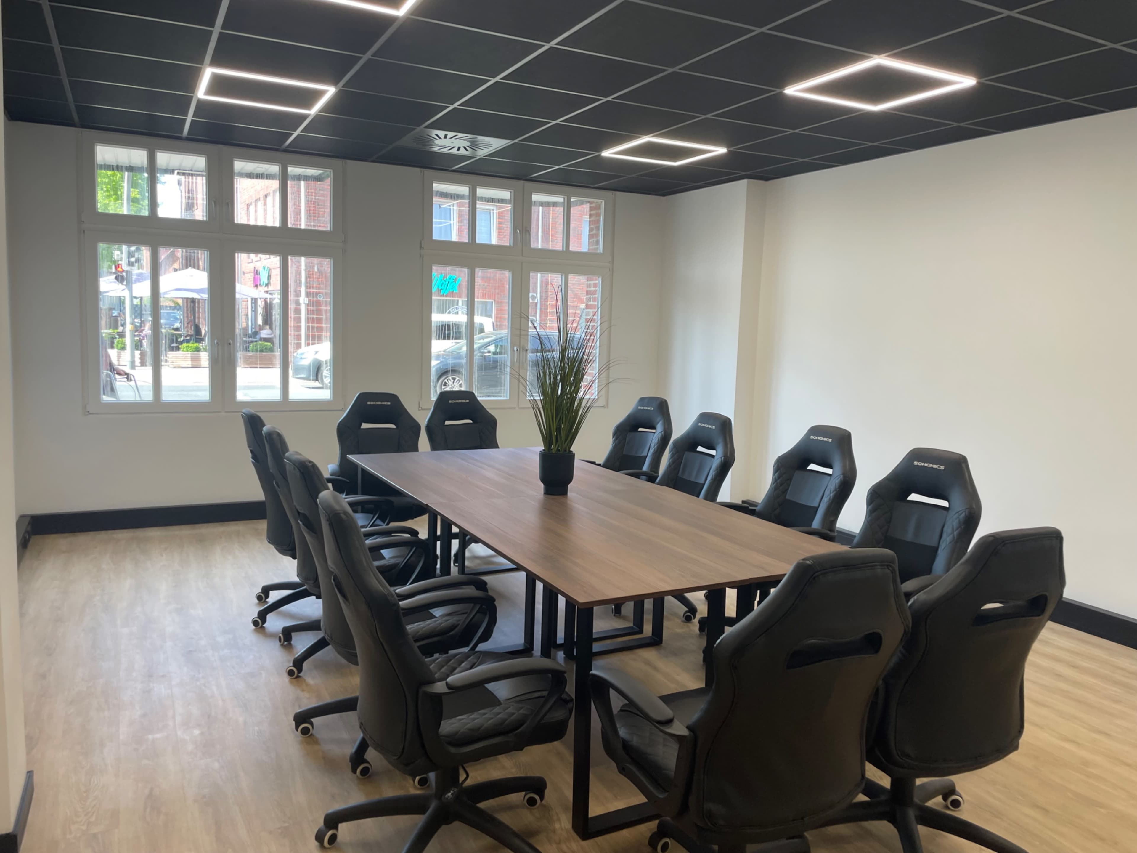 The image shows a modern conference room with a large wooden table surrounded by several ergonomic chairs, and natural light coming through multiple windows.