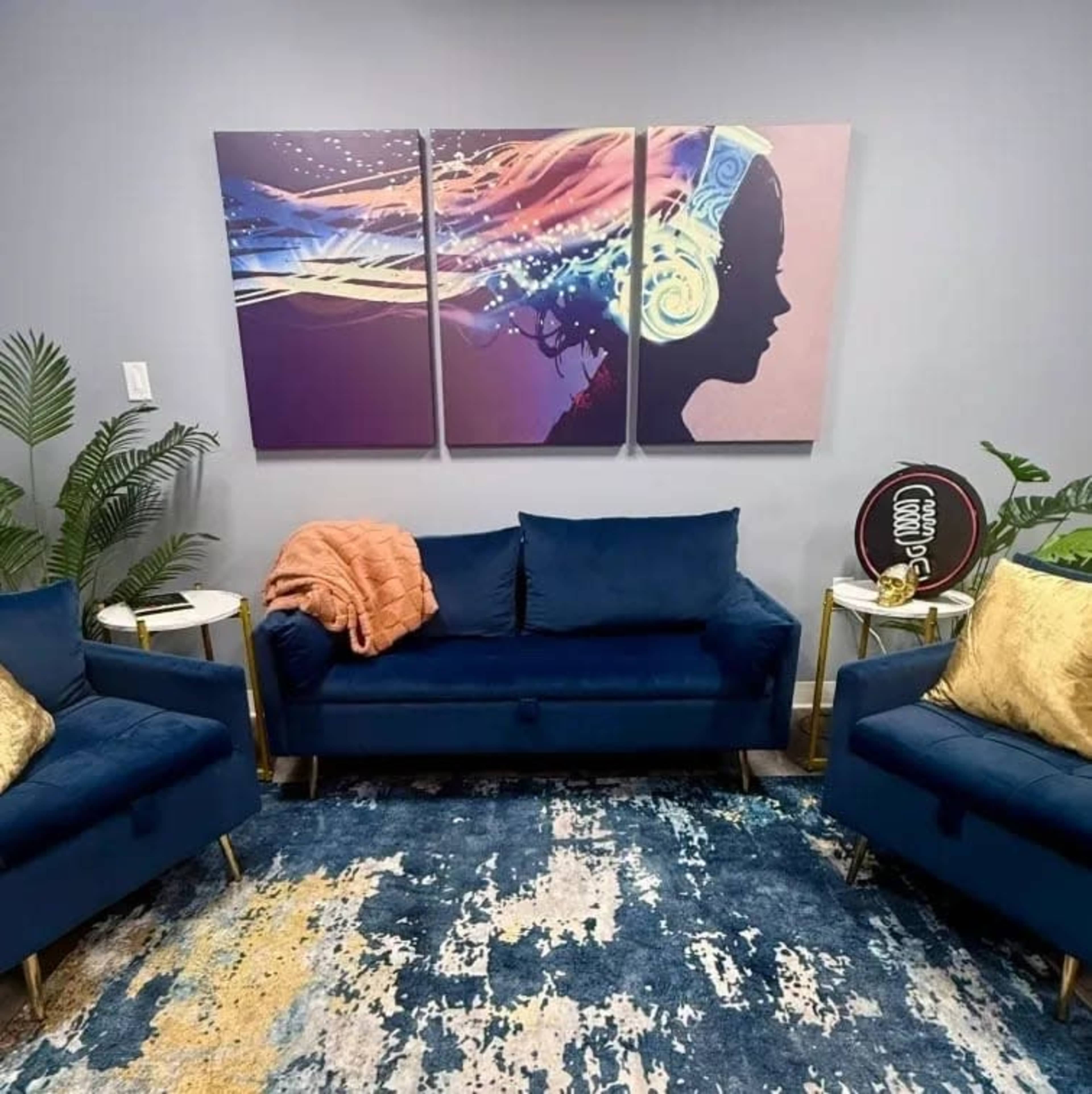The image shows a modern living room featuring a blue sofa and armchairs, a colorful wall art piece of a woman's silhouette, and decorative plants.