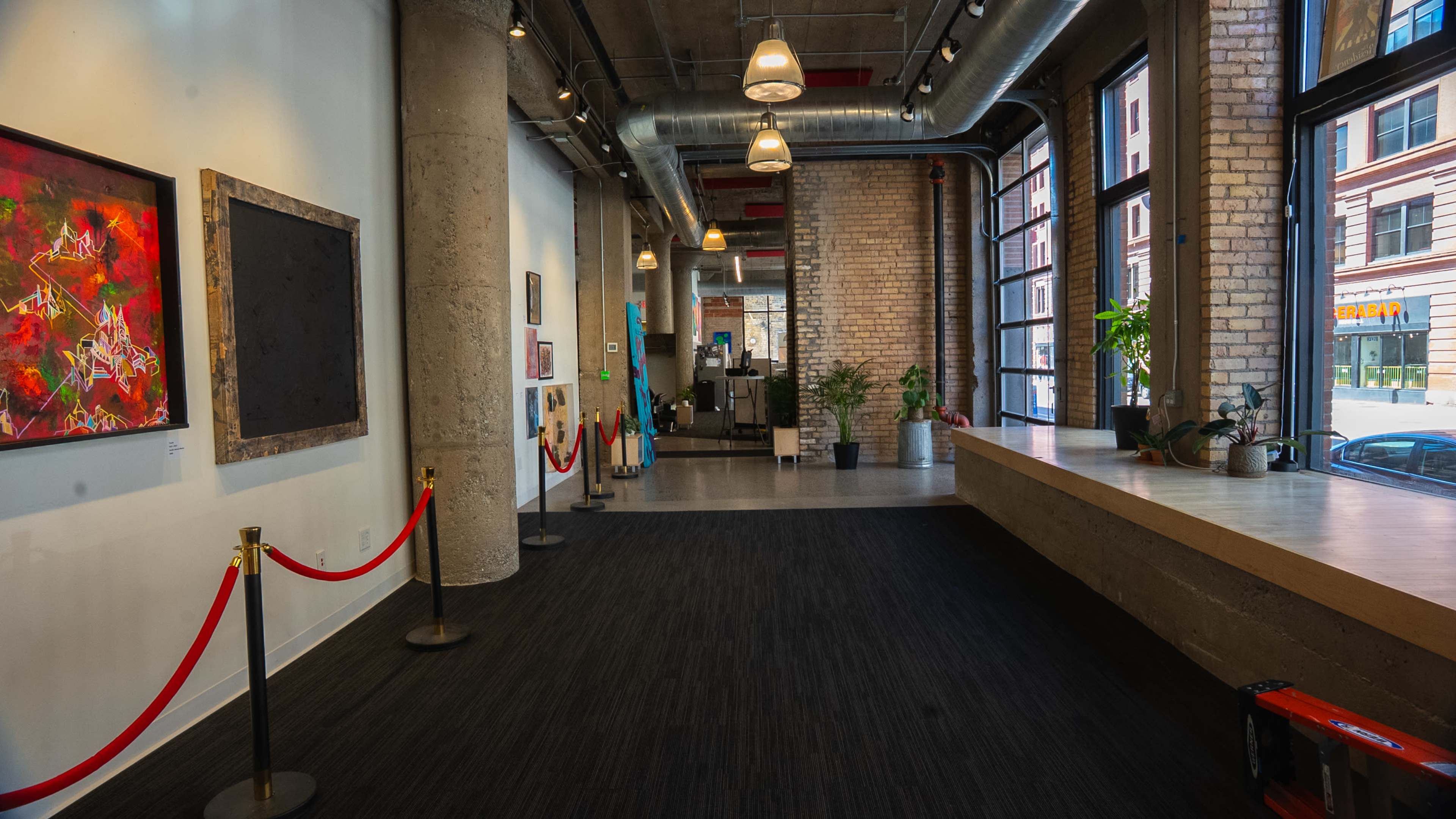 The image shows a spacious gallery or interior space featuring artwork on the walls, a red rope barrier, and large windows that overlook a street.