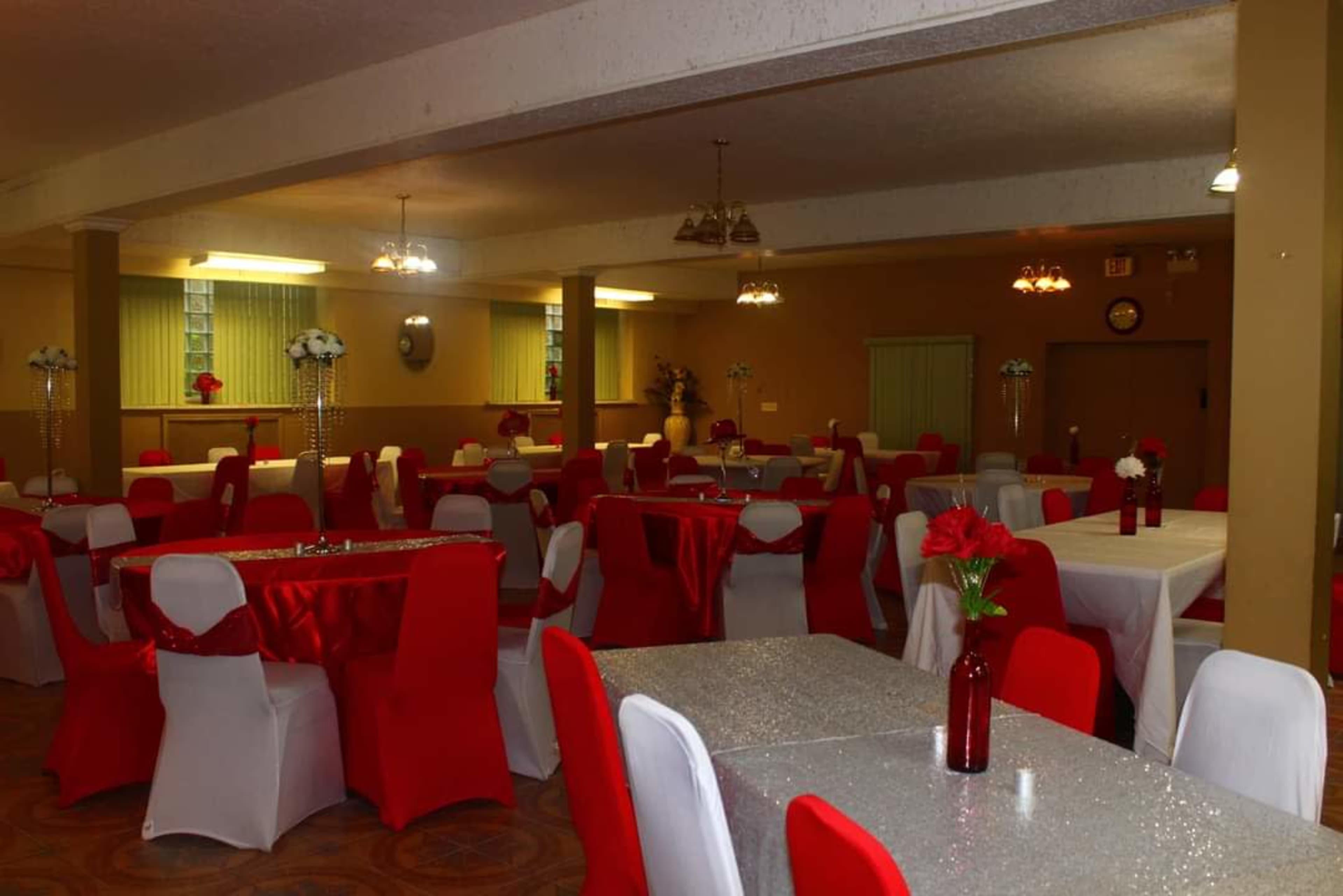 The image shows a reception hall with tables draped in white and red tablecloths, featuring red chair covers and floral centerpieces.