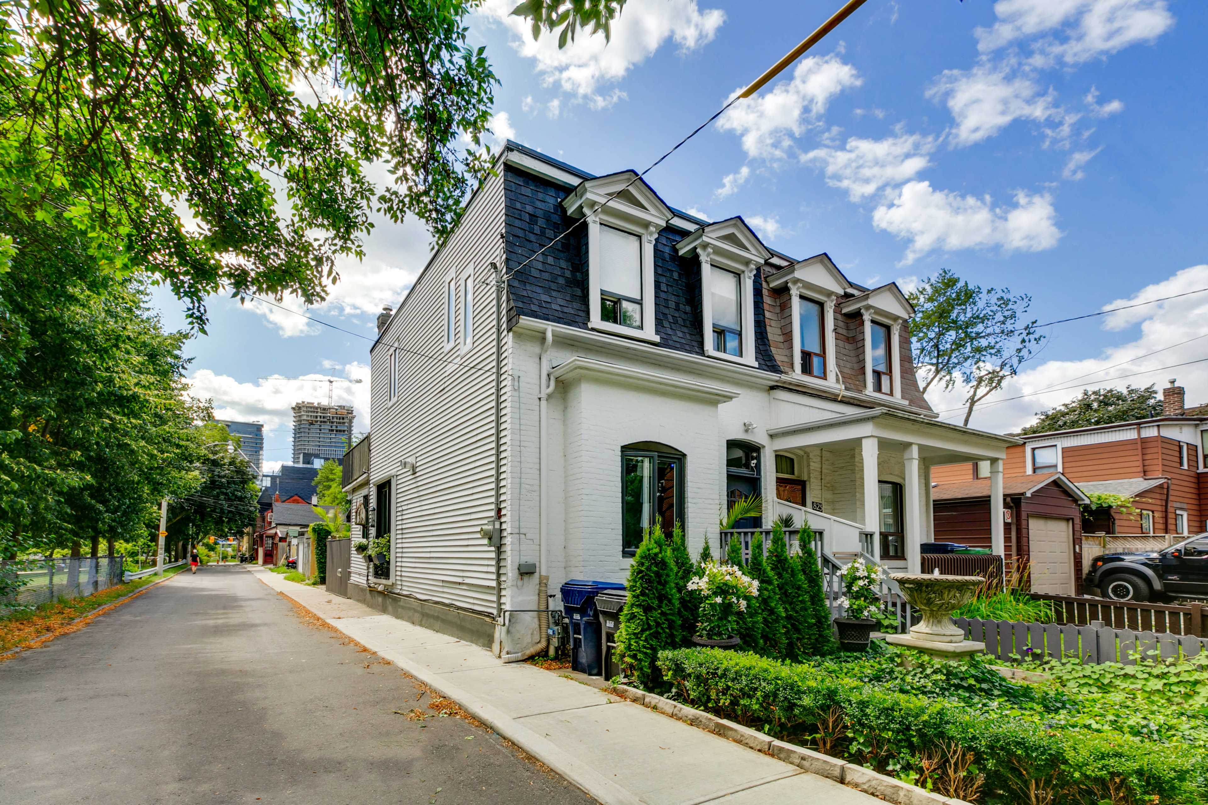 A two-story white house with a black roof located on a quiet street, surrounded by greenery and parked cars.