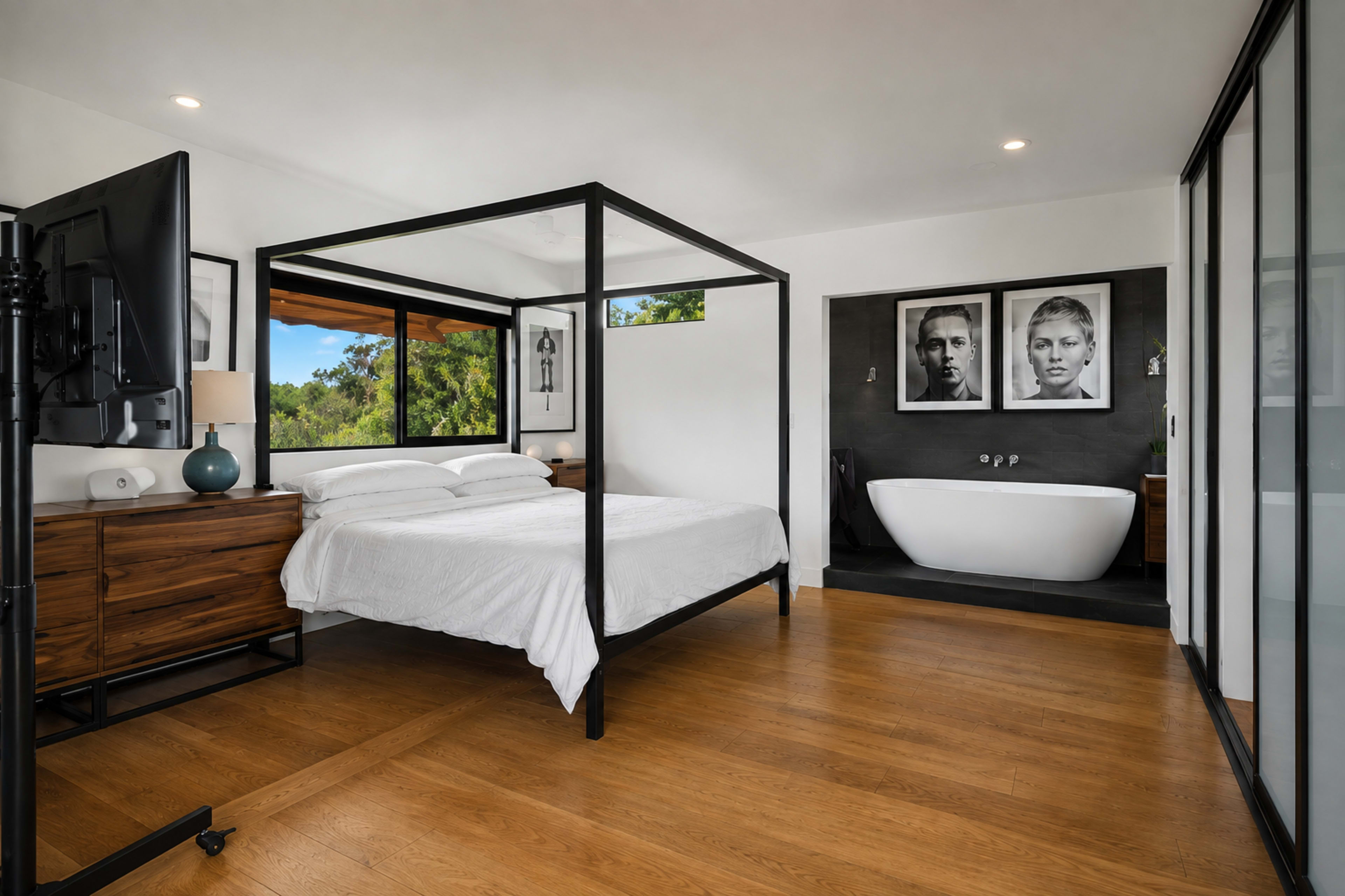 The image shows a modern bedroom with a four-poster bed, a television, large windows, and a freestanding bathtub.