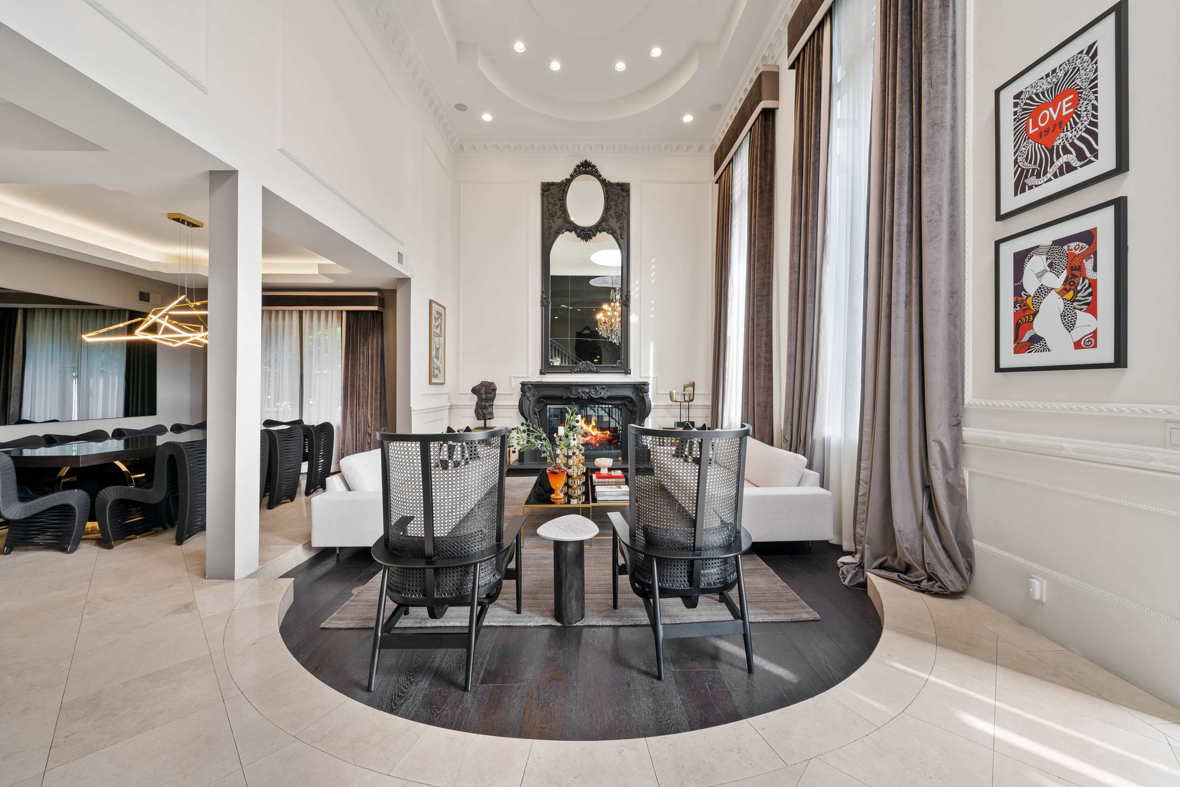 The image shows a modern living space featuring a seating area with light sofas, a dining area with black chairs, and a decorative fireplace surrounded by artwork.