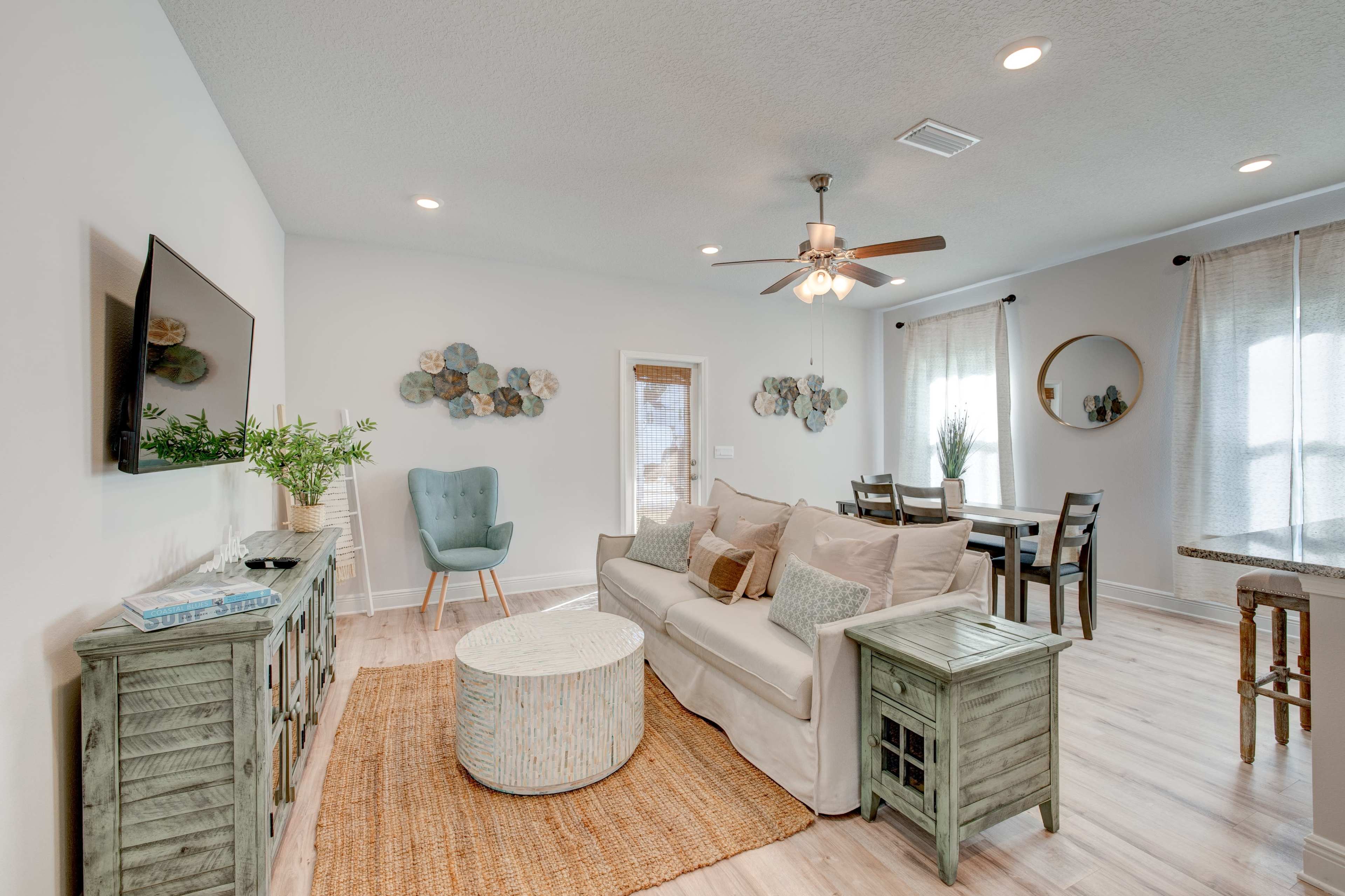 A bright living area features a light-colored sofa, a wooden coffee table, and a dining space with a round table, complemented by natural light from large windows.