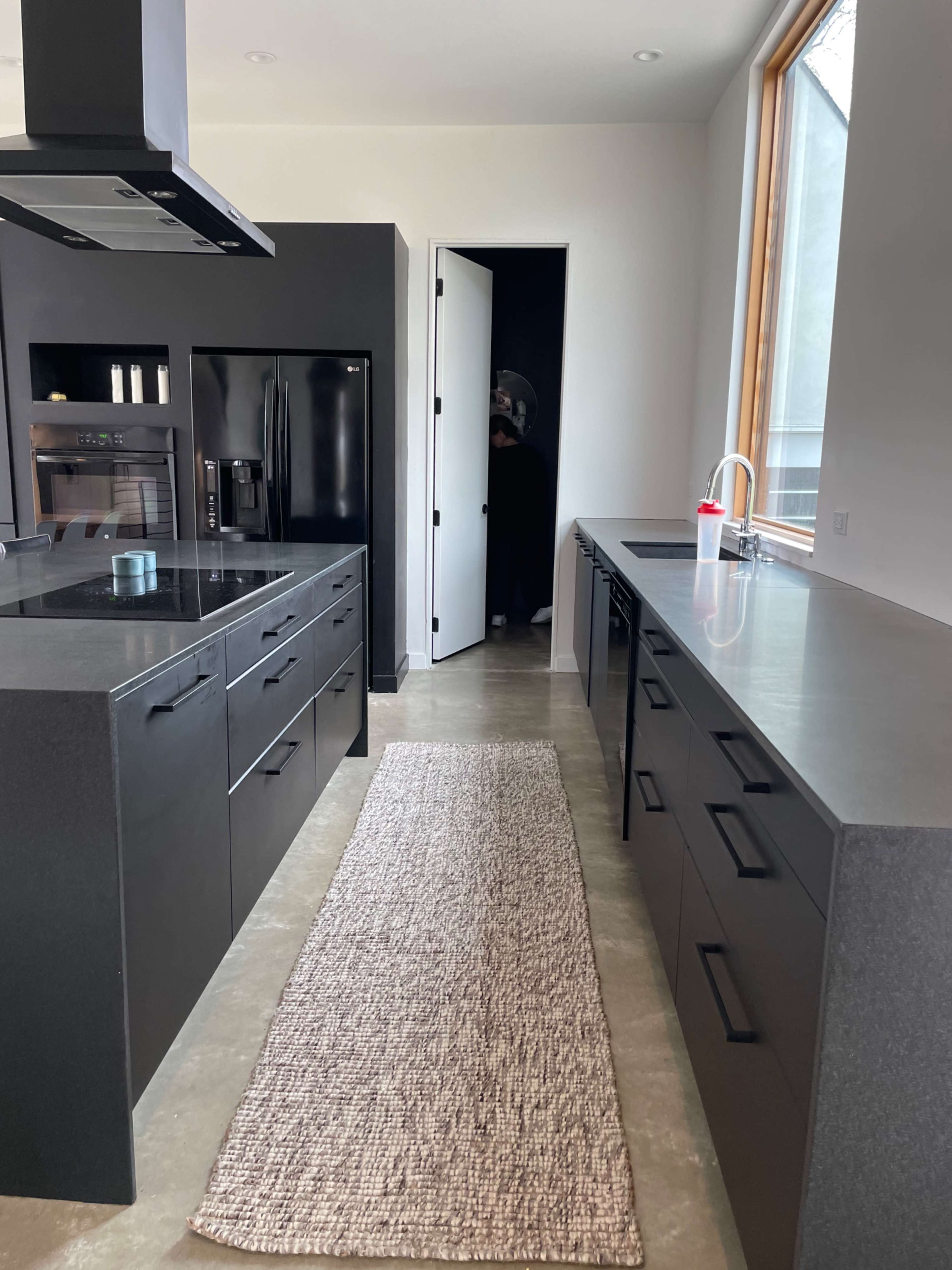 A modern kitchen features sleek black cabinetry, stainless steel appliances, and a woven runner along the concrete floor, leading to a doorway.