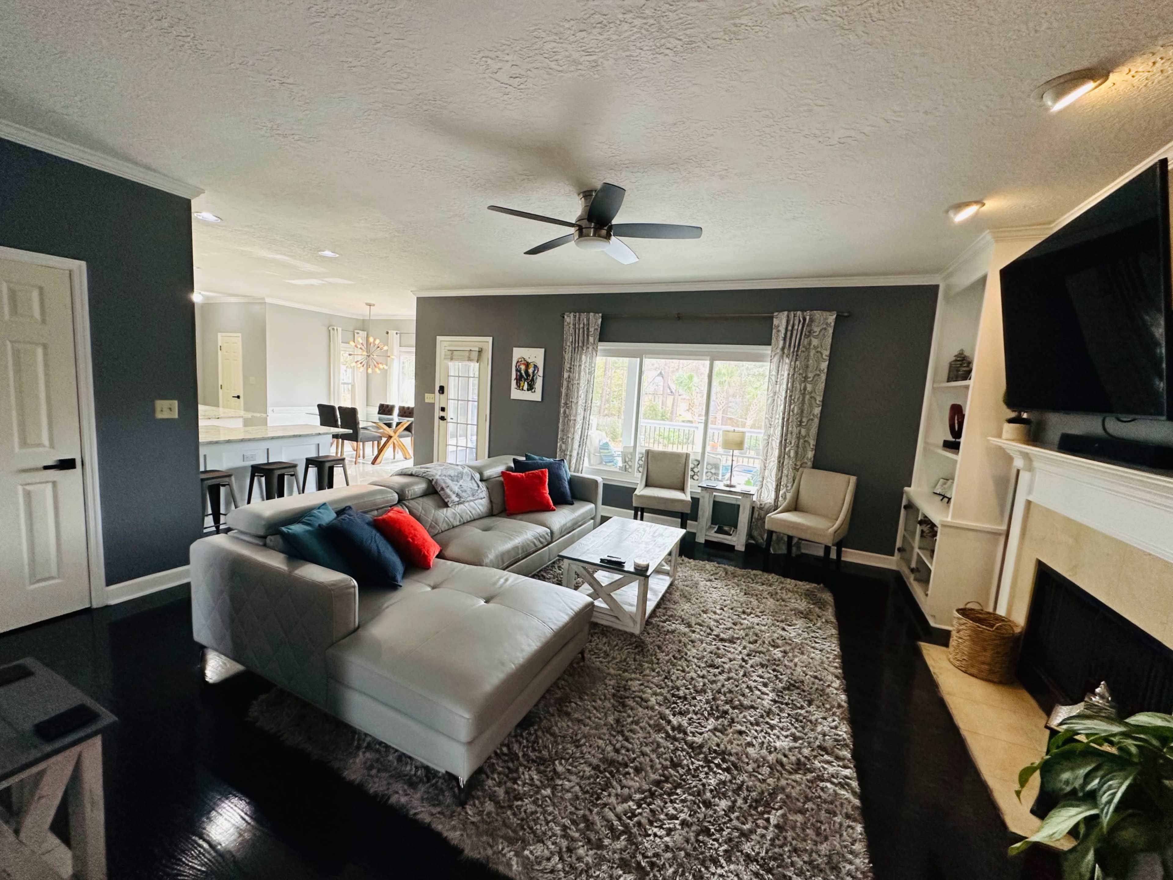 The living room features a sectional sofa with red and blue pillows, a large area rug, and a window with natural light, complemented by a ceiling fan and a fireplace.