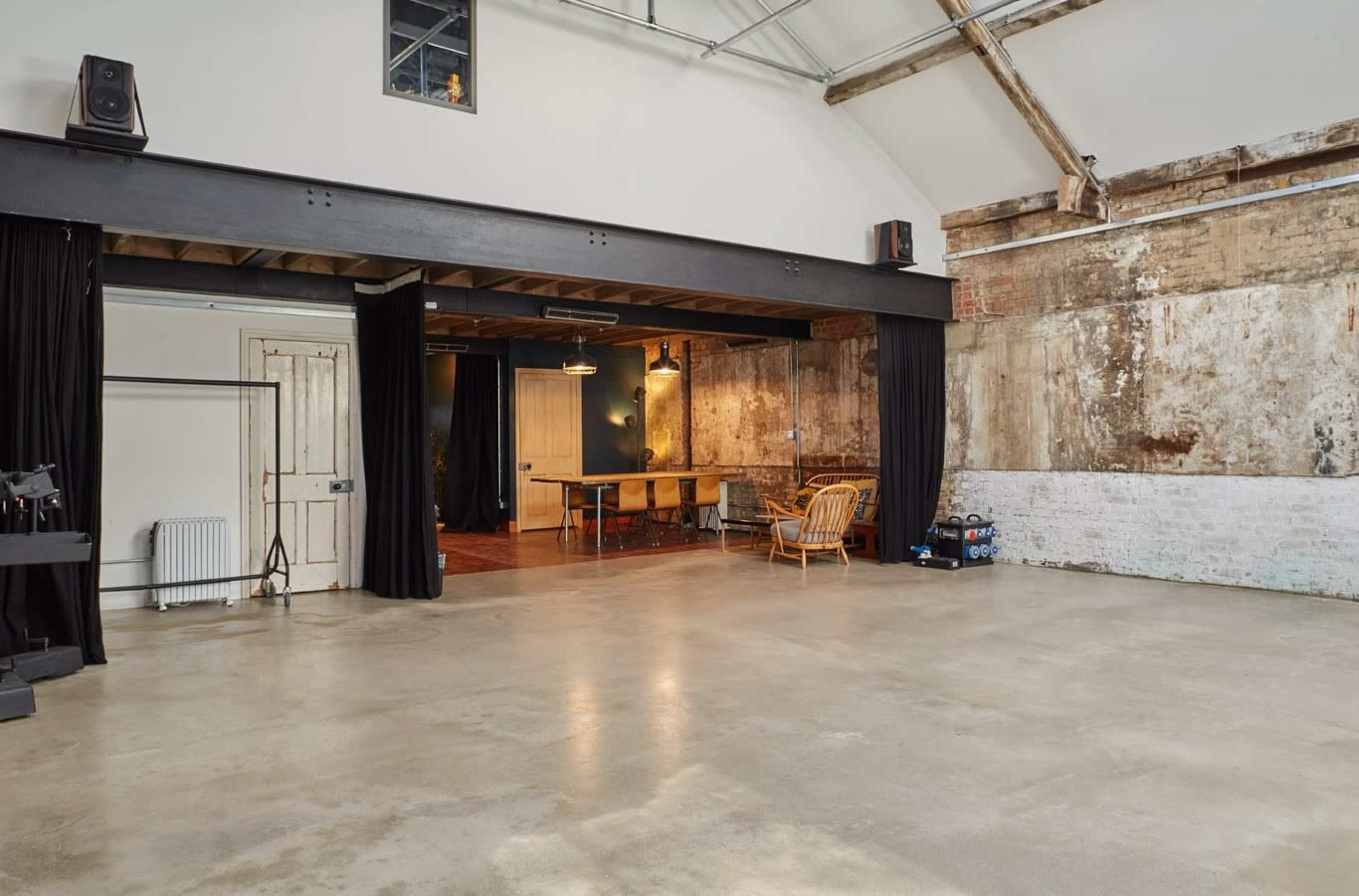 The image shows a spacious room with a polished concrete floor, exposed brick walls, wooden beams, and curtains leading to a separate area furnished with a table and chairs.