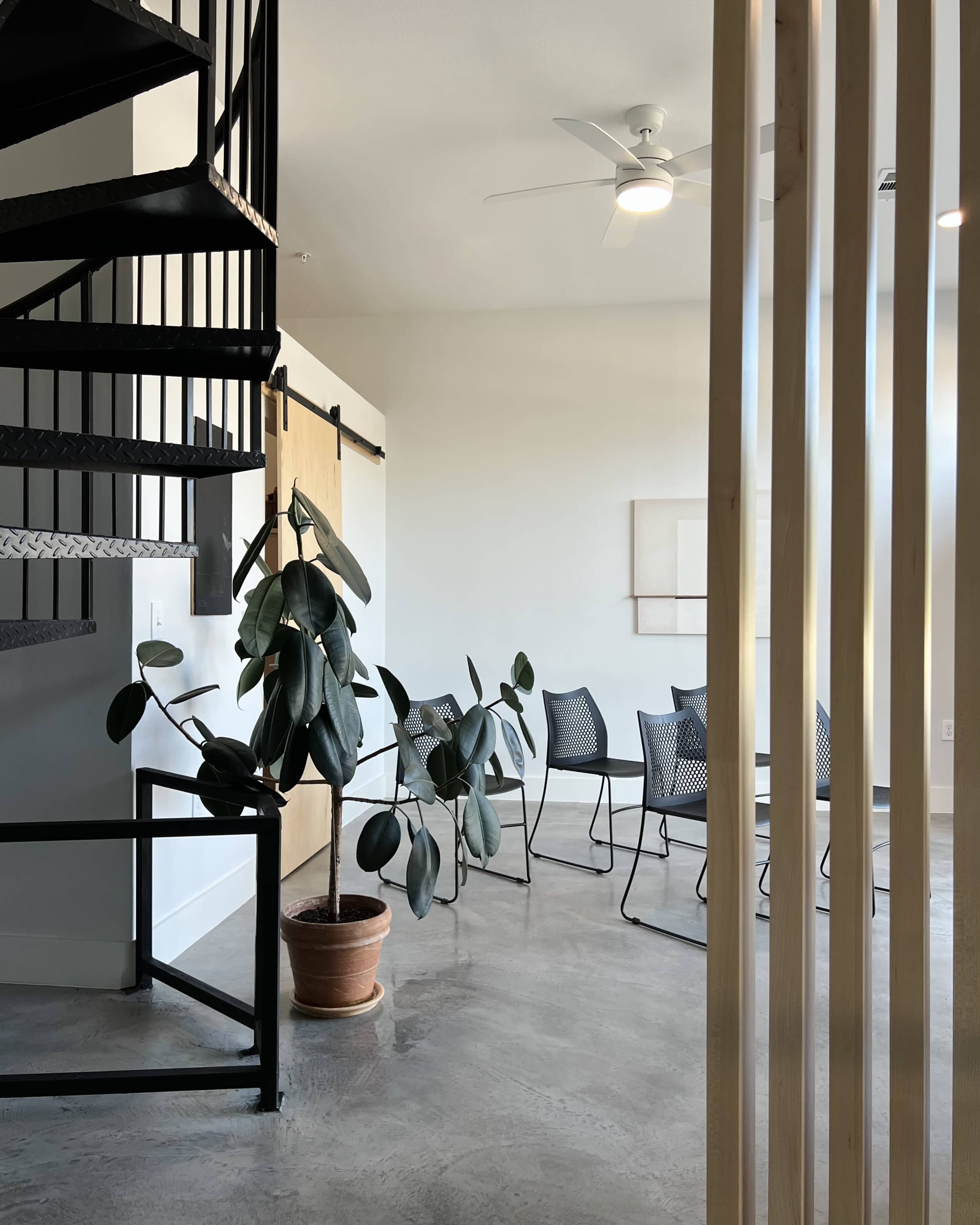 The image shows a modern interior space with a spiral staircase, a potted plant, and metal chairs arranged around a table.