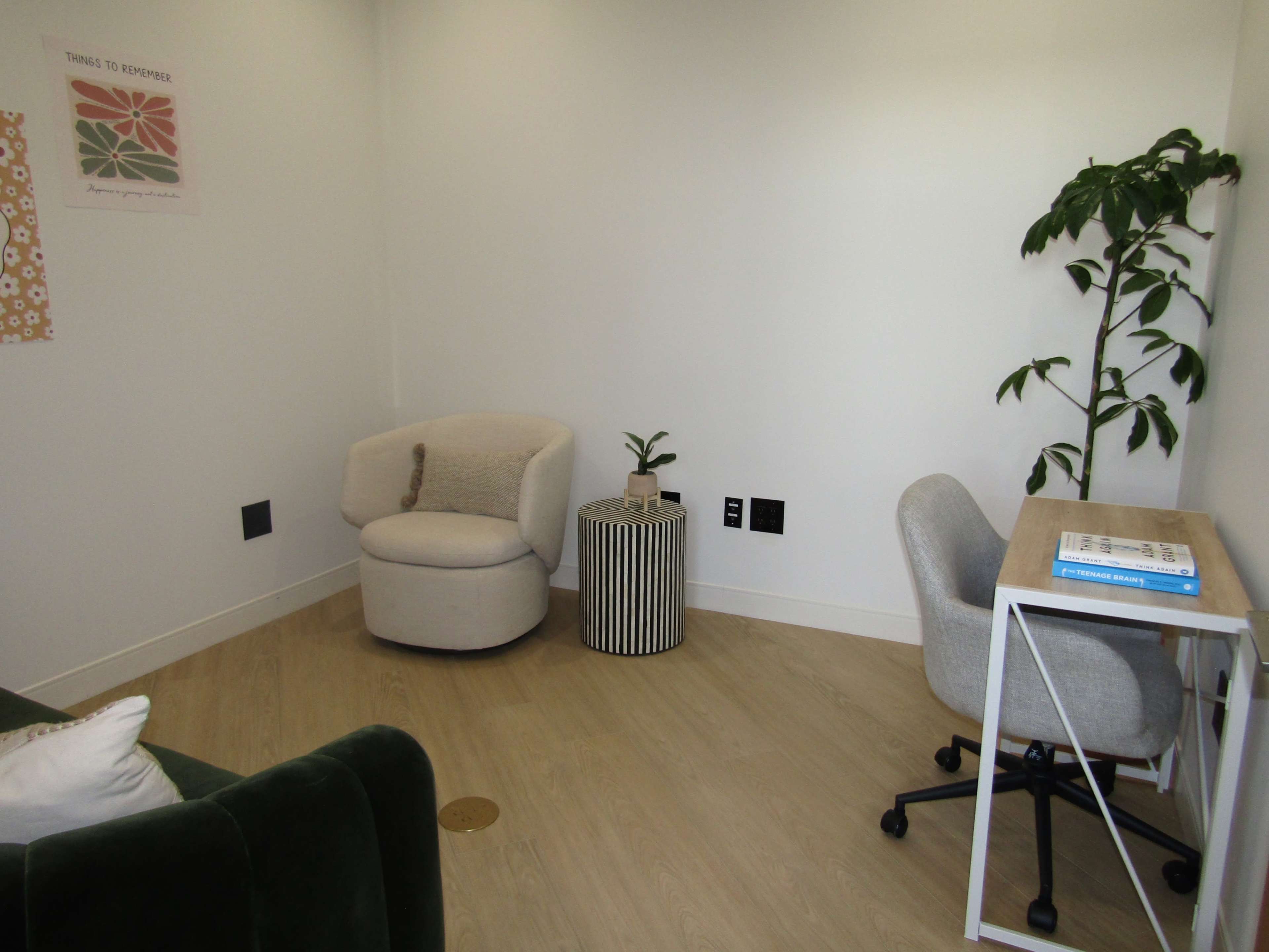 The image shows a minimalist room with a circular chair, a small plant on a cylindrical table, and a desk with a gray chair against a white wall.