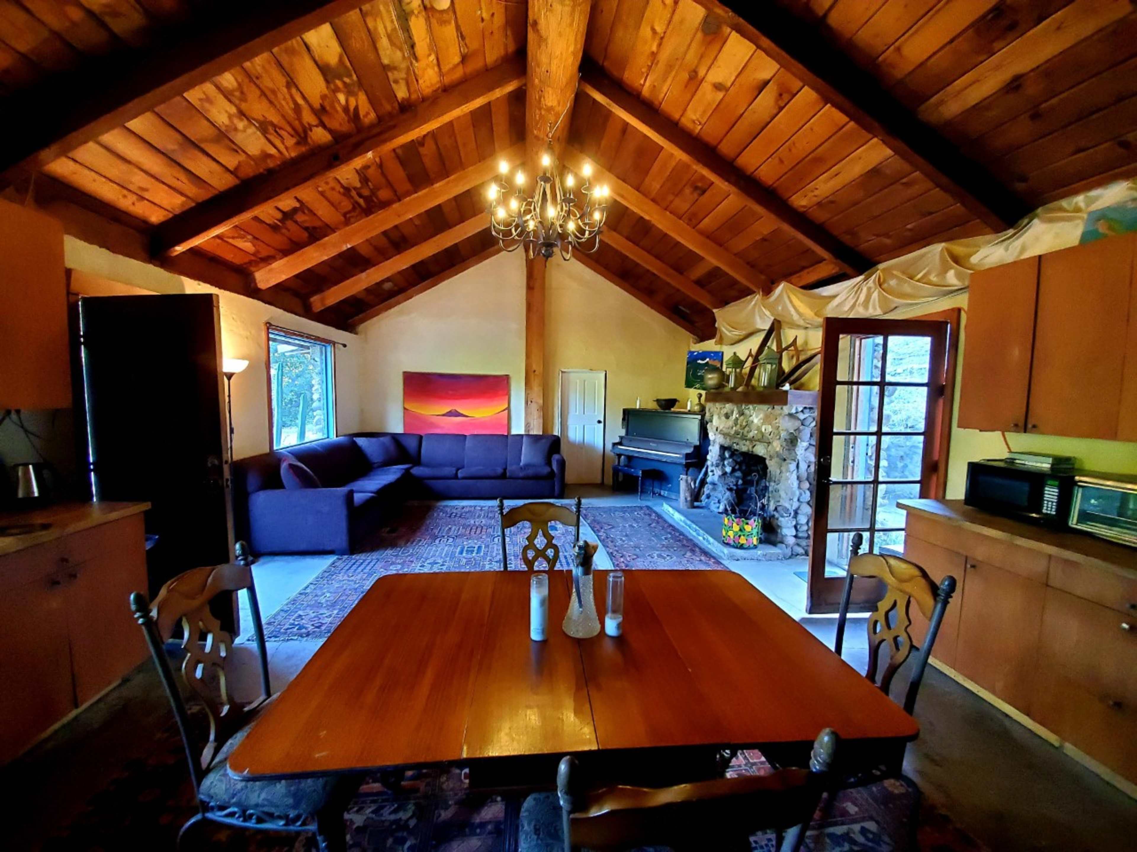 A spacious interior of a cabin features a dining table, a large couch, a grand piano, and a stone fireplace under a wooden ceiling with exposed beams.