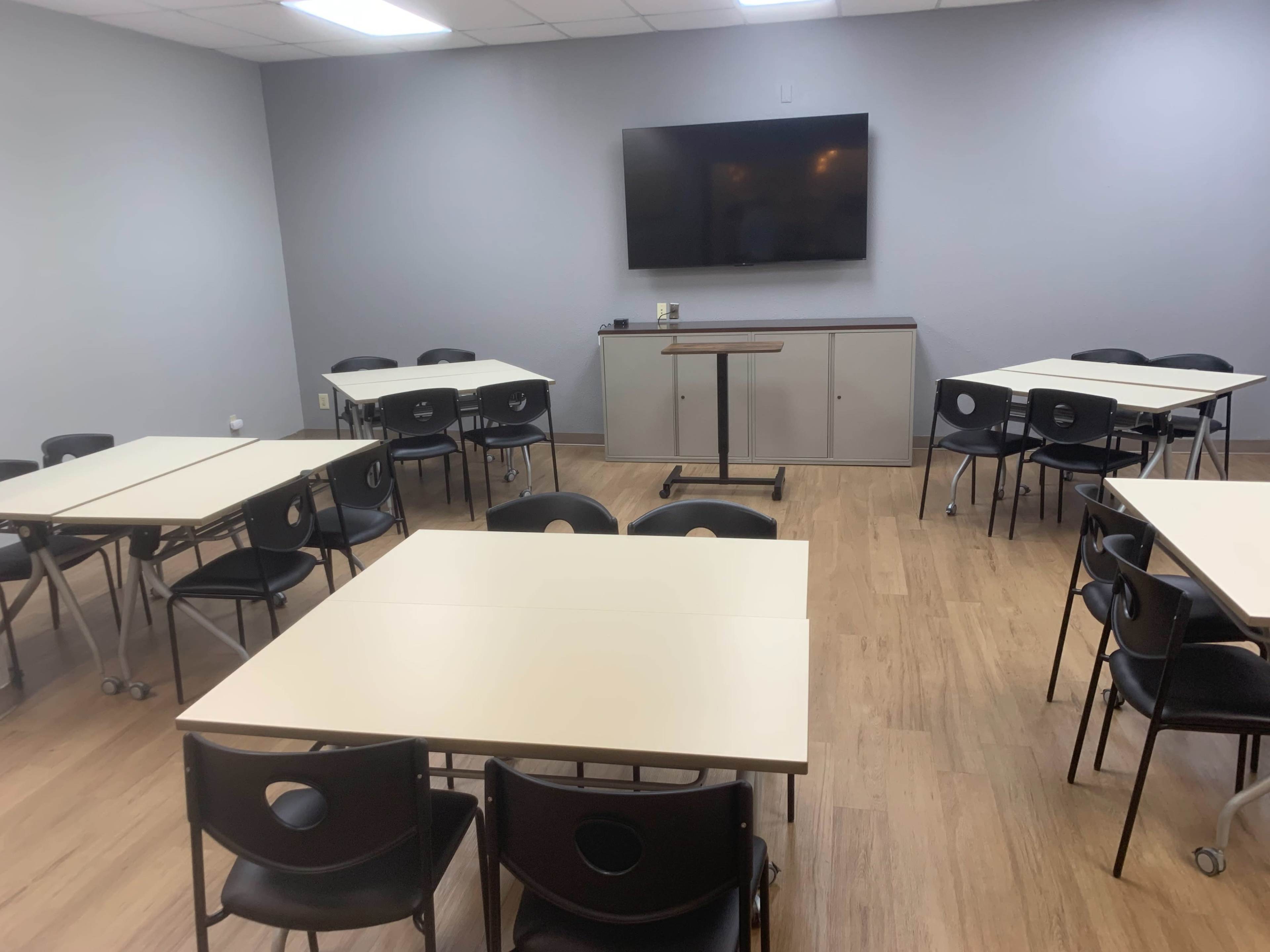 A classroom is arranged with several movable tables and chairs, featuring a wall-mounted television at the front.