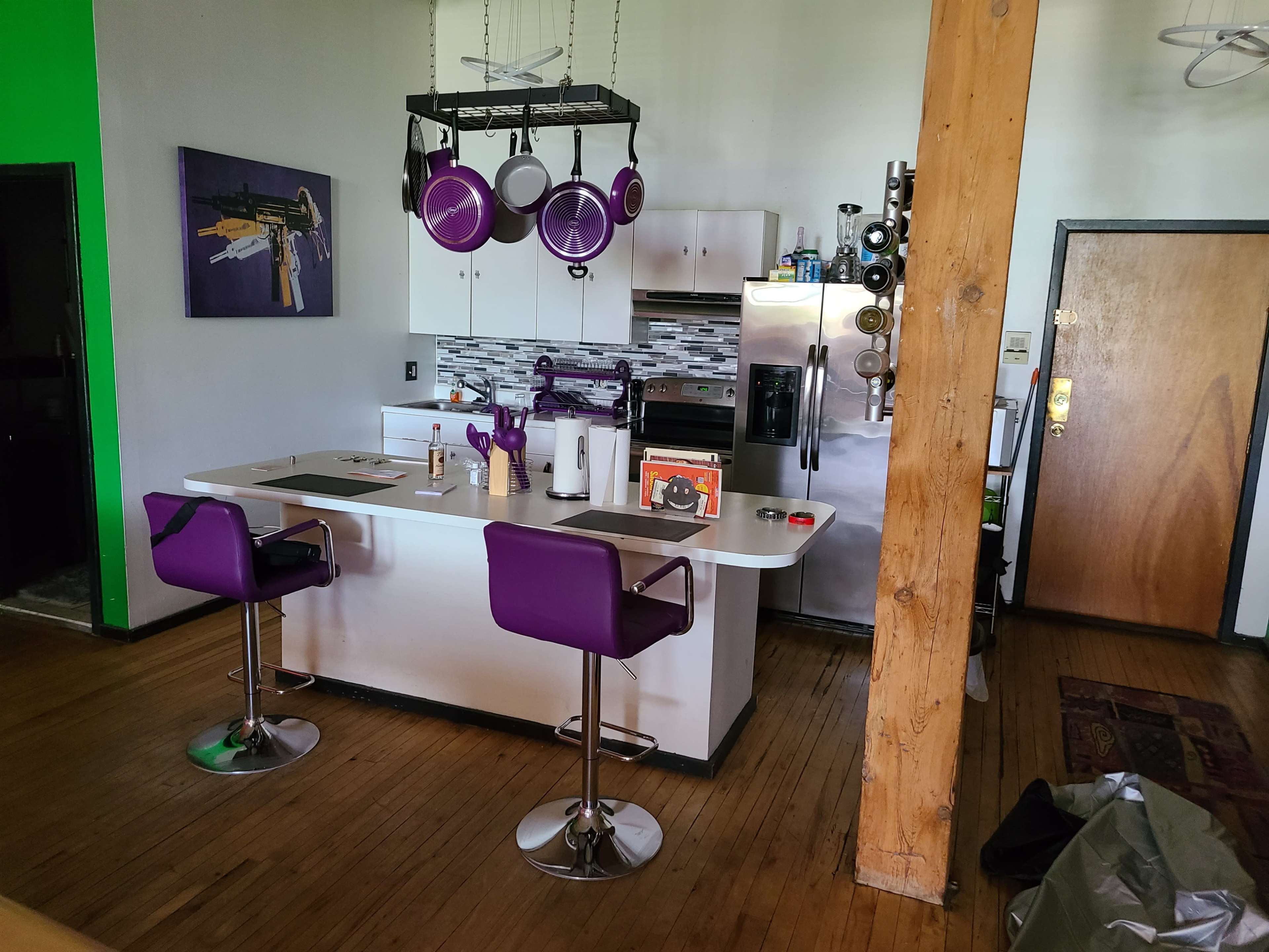 A modern kitchen with purple bar stools, stainless steel appliances, and a large island featuring cooking utensils and a decorative plant.