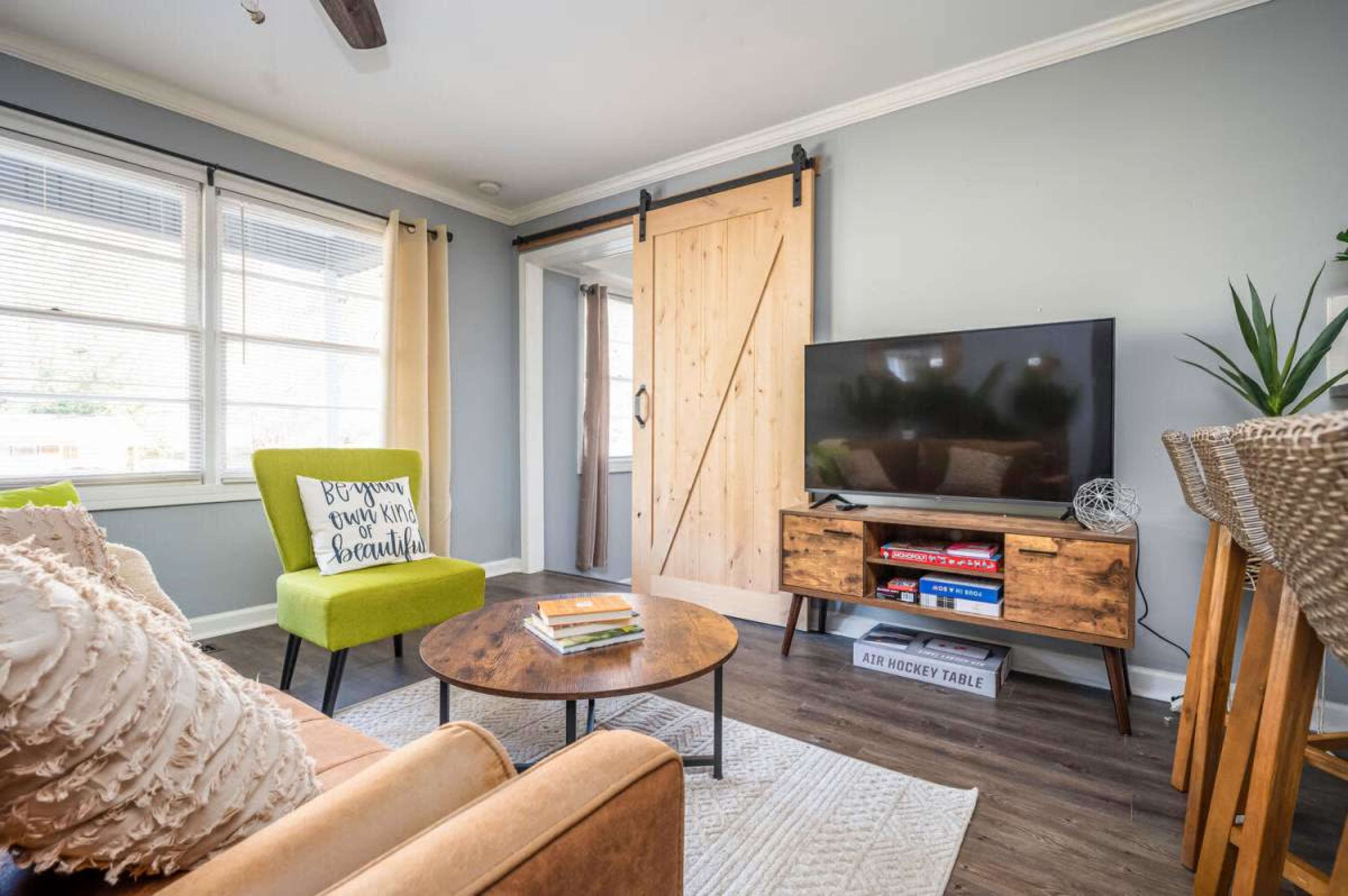 The image shows a cozy living room featuring a green accent chair, a wooden television stand, and a sliding barn door.