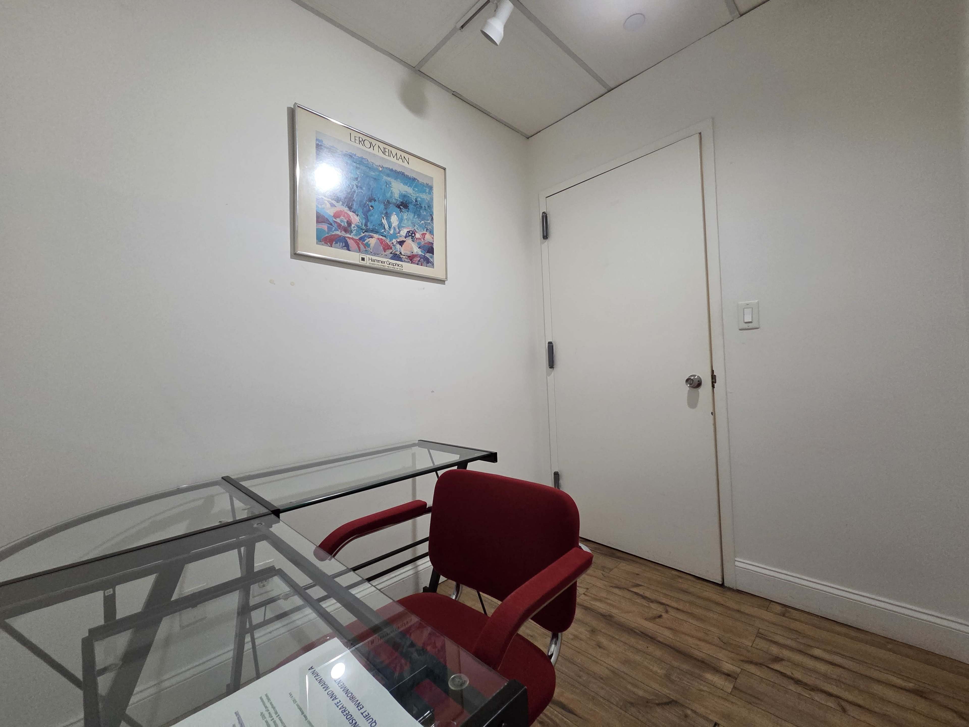 The image shows a small room with a glass desk, a red chair, a framed picture on the wall, and a closed door.