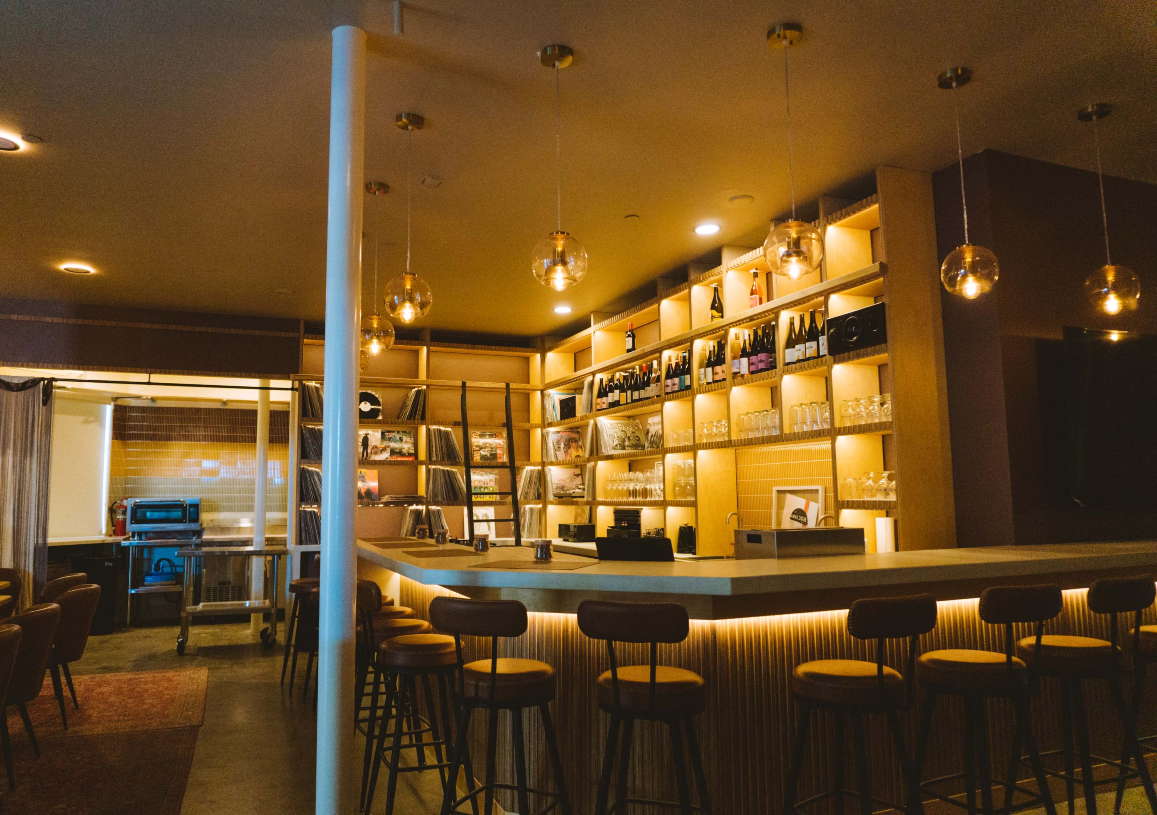 The image shows a modern bar area with a well-lit counter, shelves displaying bottles of wine and glassware, and stylish pendant lighting hanging overhead.