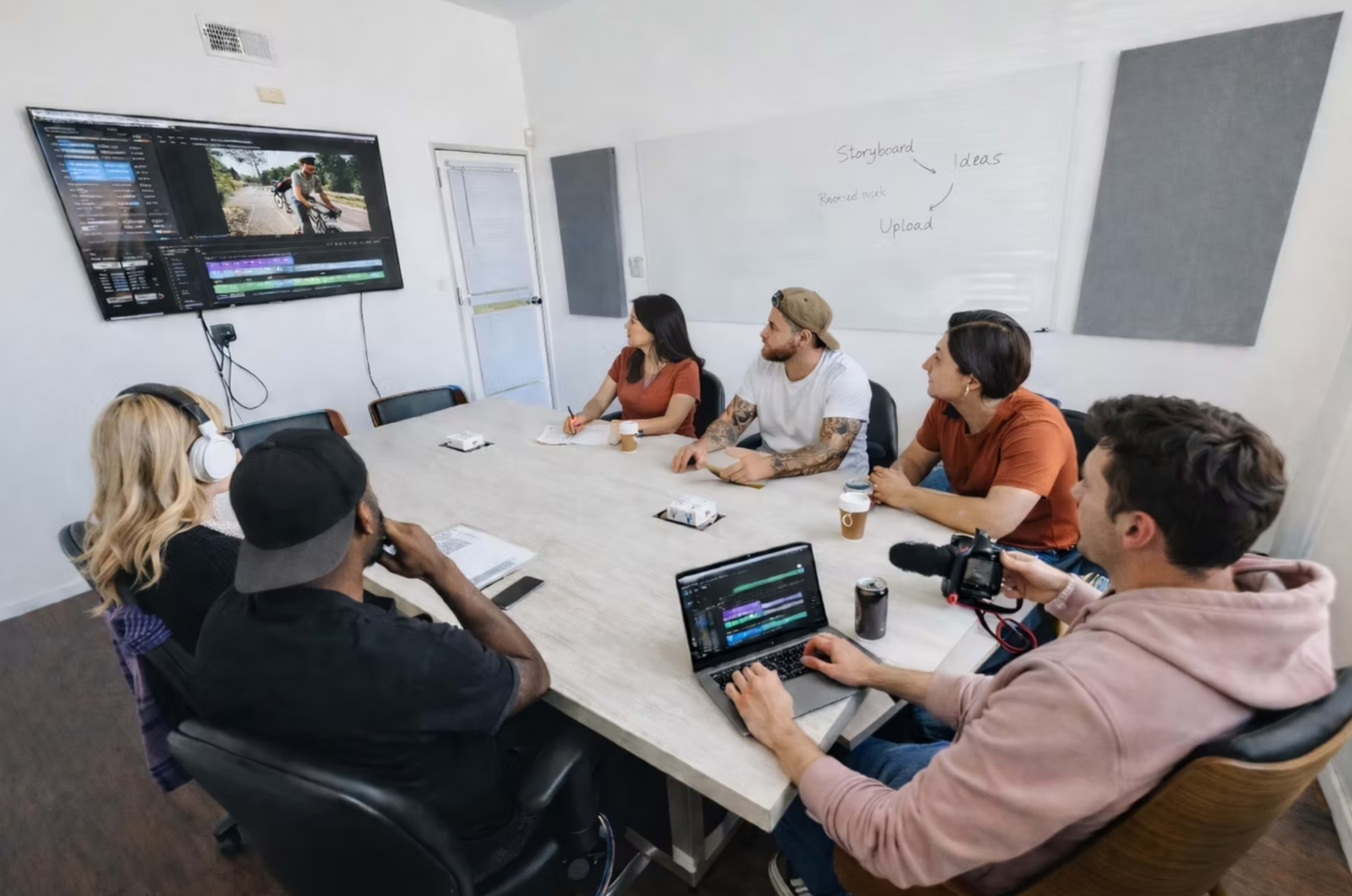 A group of six people sits around a large table in a bright room, watching a video editing presentation on a screen while taking notes.