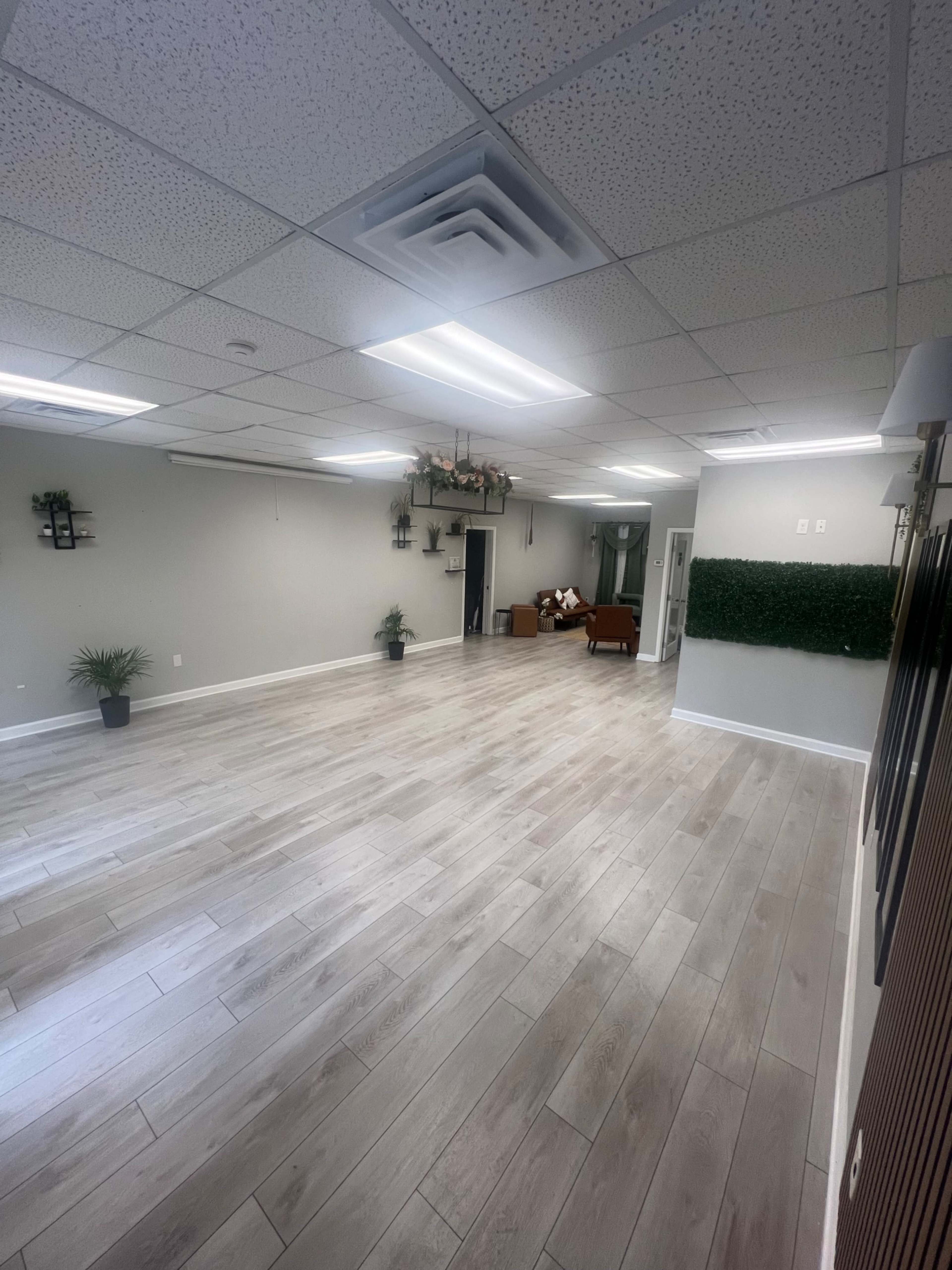 The image shows a spacious, empty room with light-colored wooden flooring, green plants along the walls, and a seating area in the background.