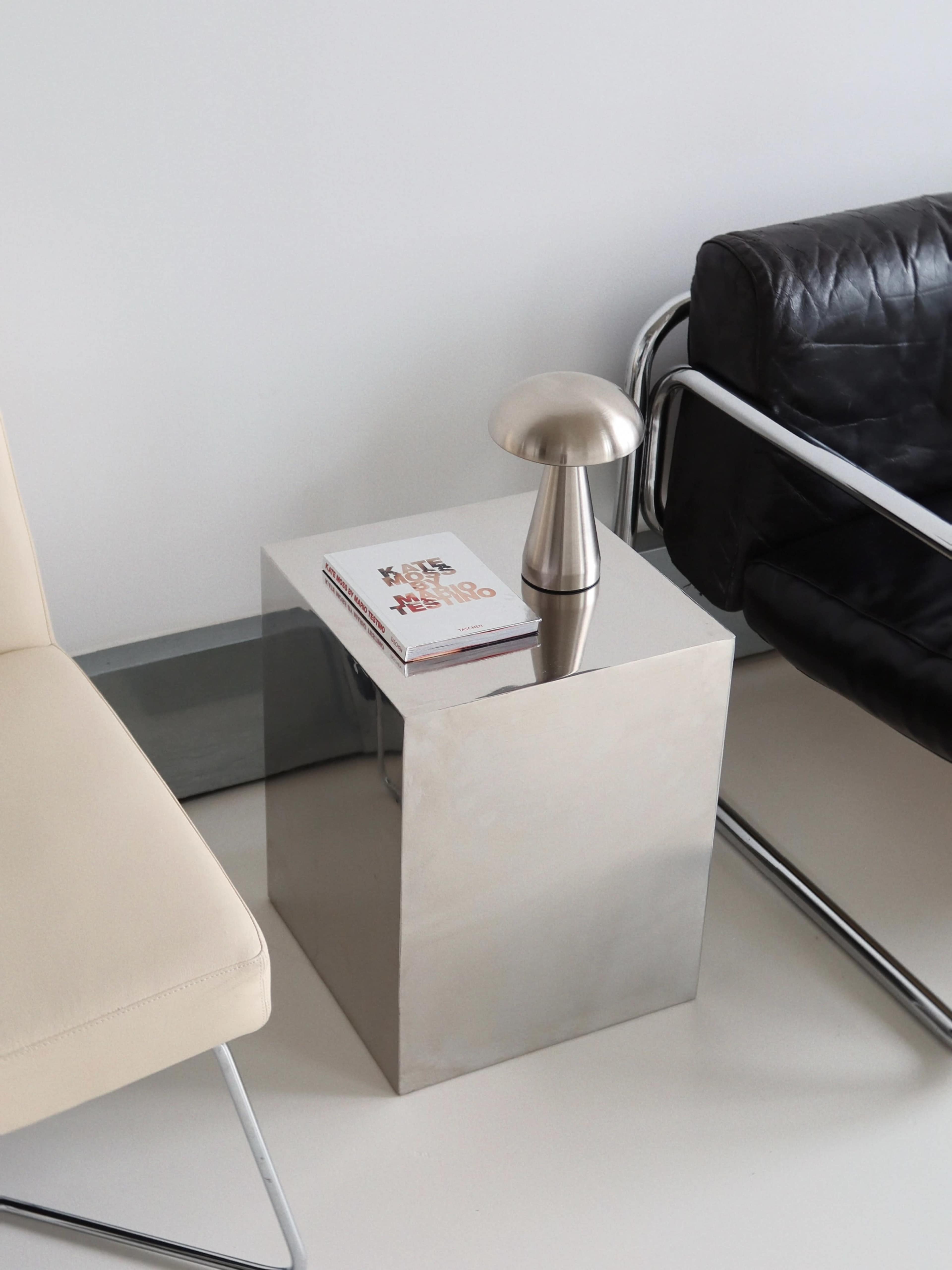 A metallic side table holds a lamp and a stack of books between a black chair and a beige chair.