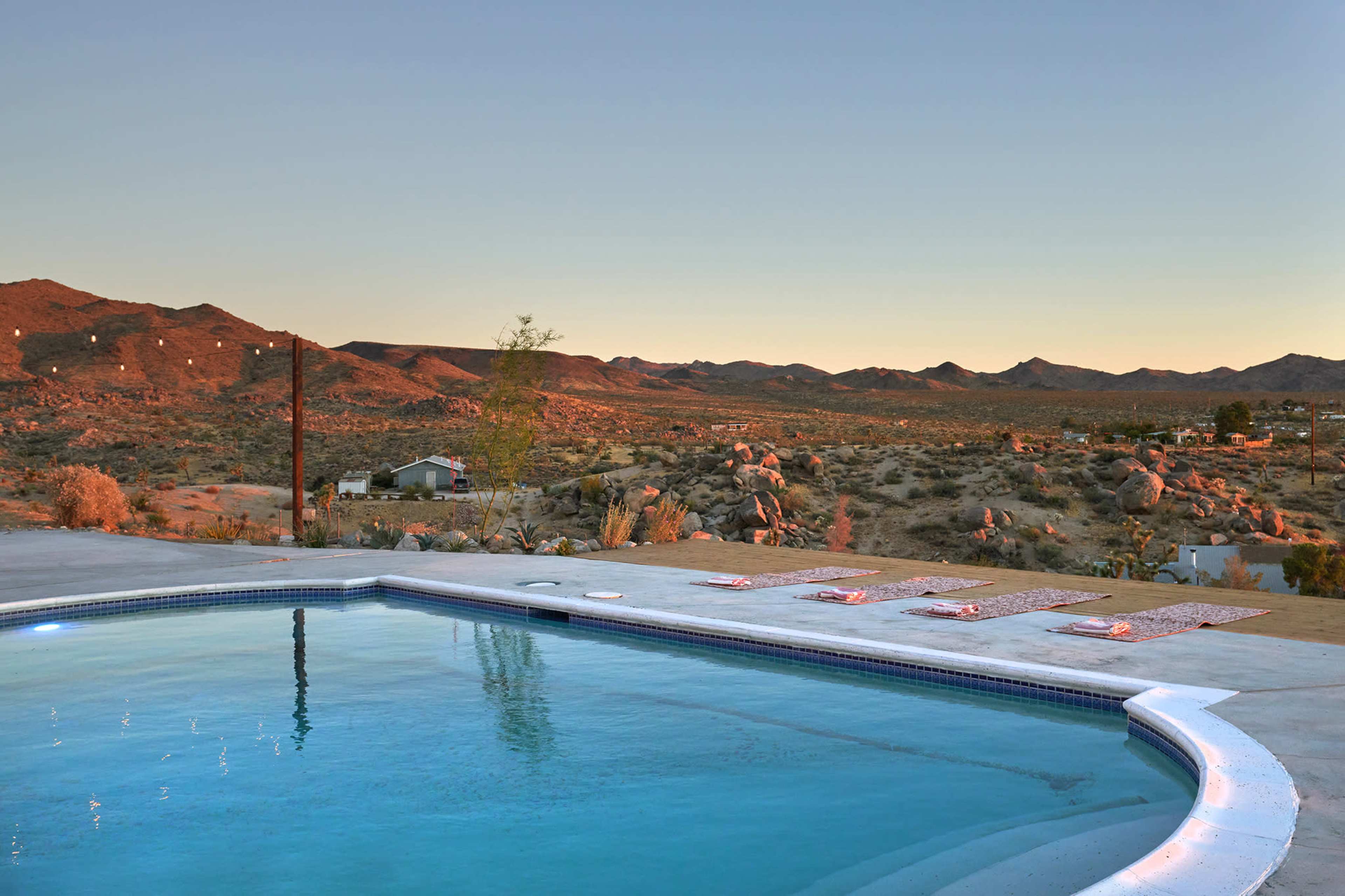 A swimming pool is situated in the foreground, overlooking a desert landscape with mountains and sparse vegetation at sunset.