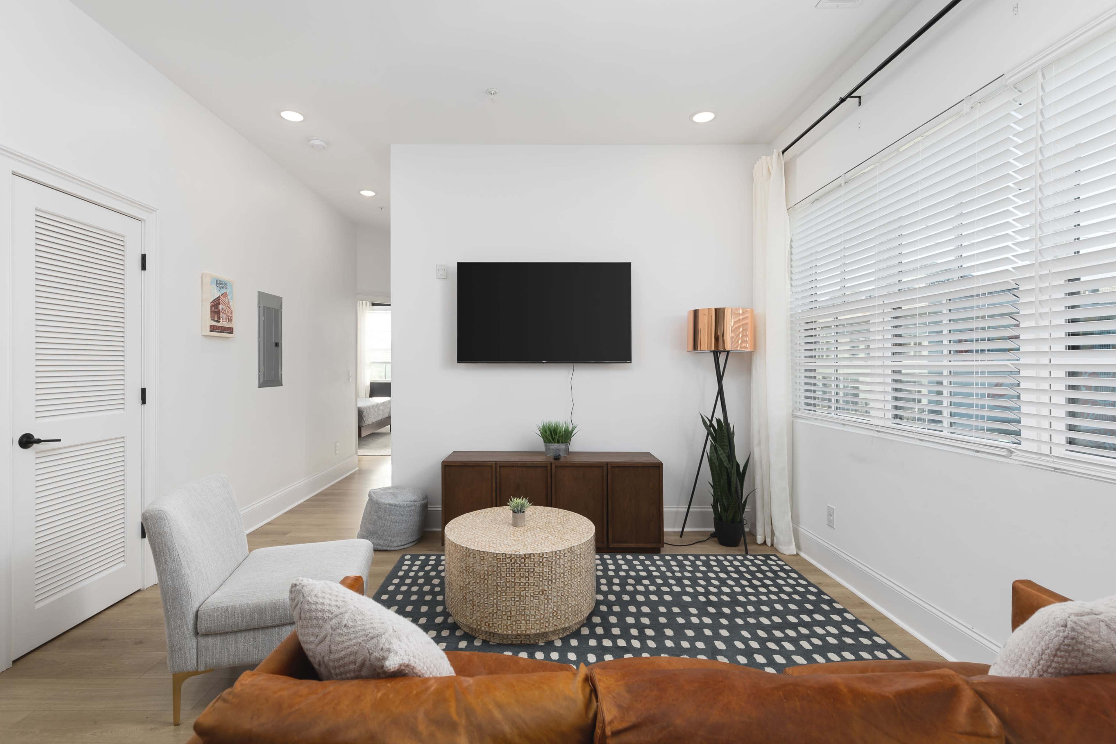 The image shows a modern living room with a brown leather sofa, a round coffee table, a television mounted on the wall, and plants in the corner, all featuring a light color scheme.