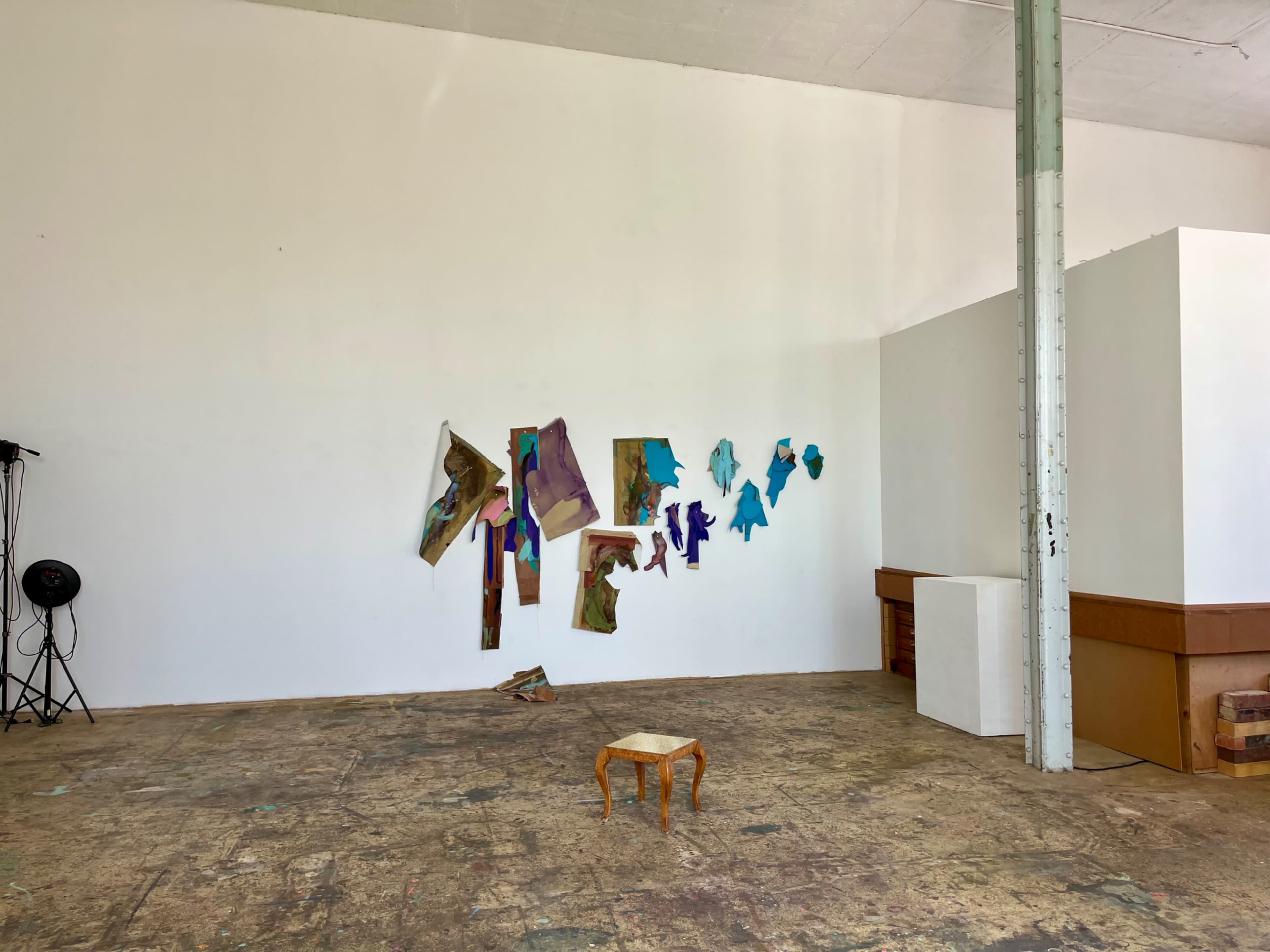 The image shows a spacious, minimally furnished room with a small wooden stool in the center and colorful art pieces attached to a white wall.