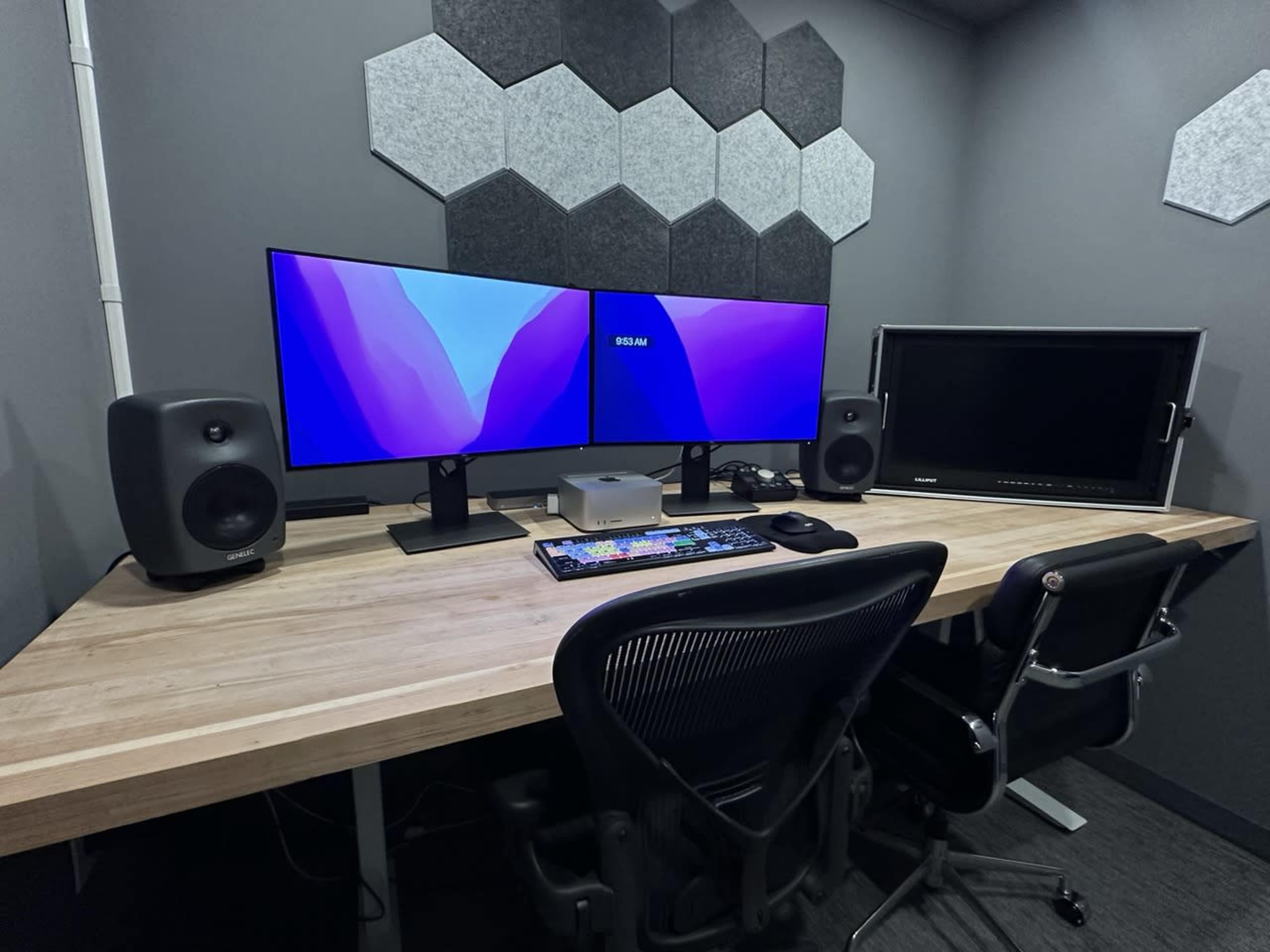 A modern workspace featuring two large monitors, a desktop computer, speakers, and a wooden desk with two office chairs.