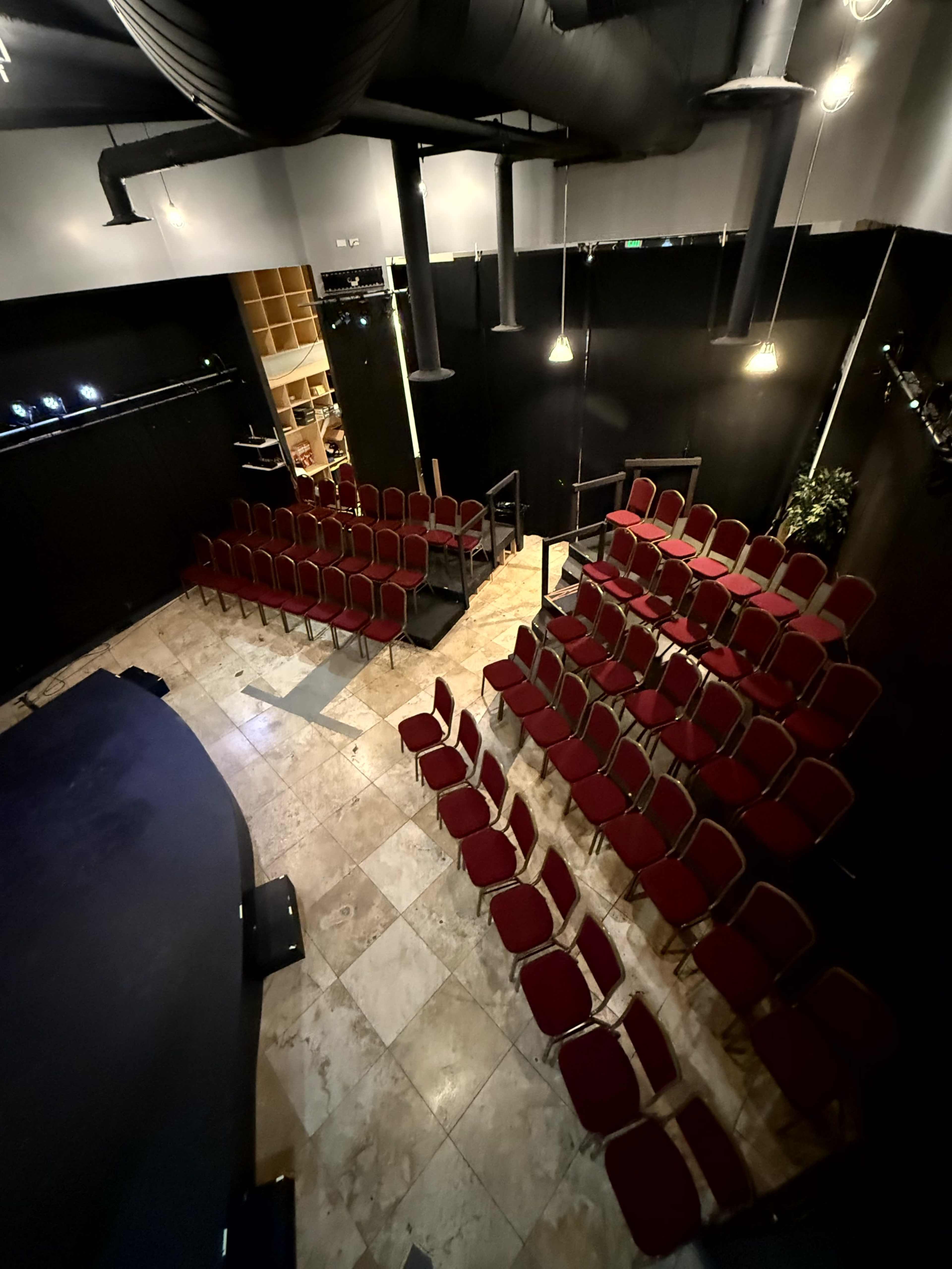 The image shows an indoor theater with a tiered seating arrangement of red chairs facing a small stage.