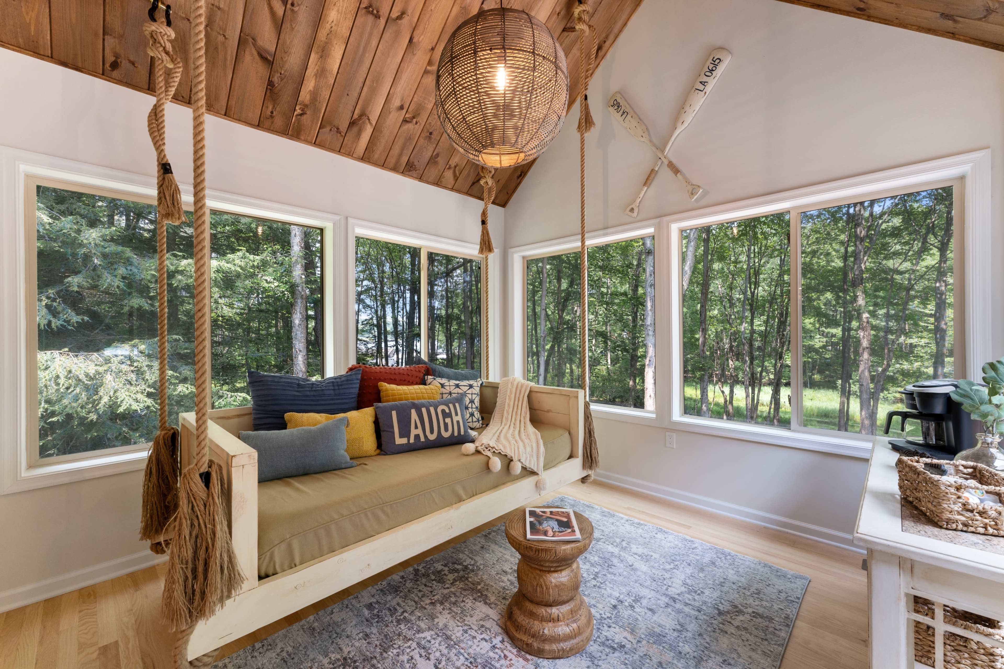 The image shows a cozy sitting area with a swing-style bench, large windows overlooking trees, and a small table featuring decorative items.