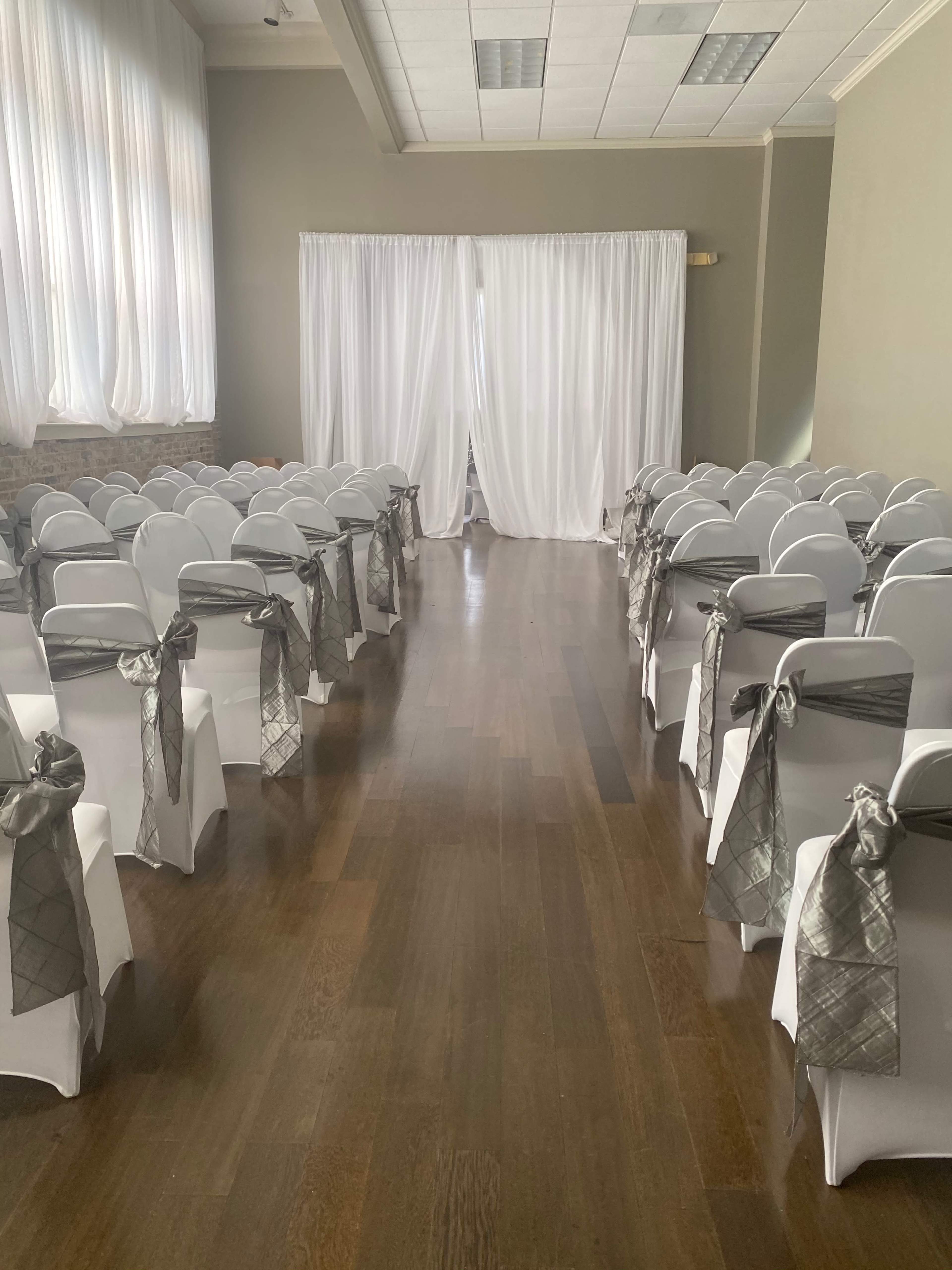 A wedding ceremony setup with white chairs arranged in rows, each adorned with silver sashes, leading toward a white draped backdrop.