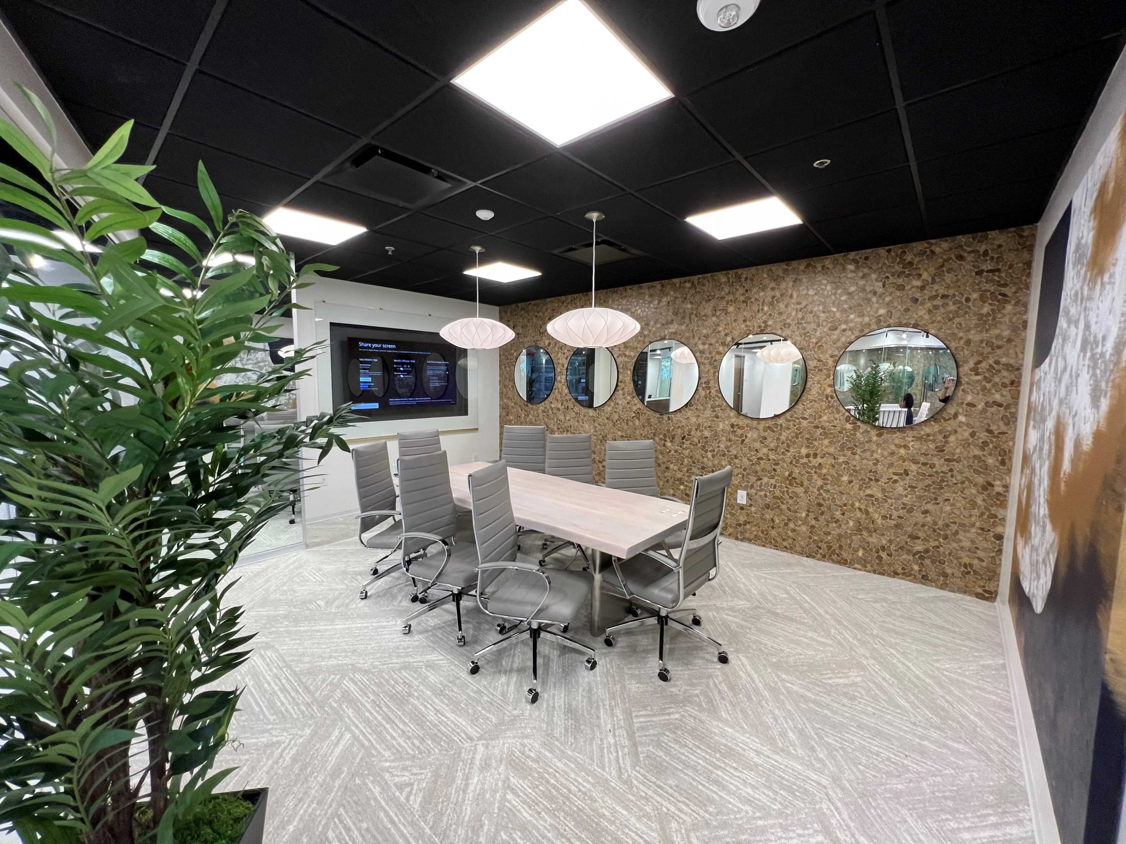 A modern conference room features a large table surrounded by chairs, with a wall of mirrors and a screen displaying information.