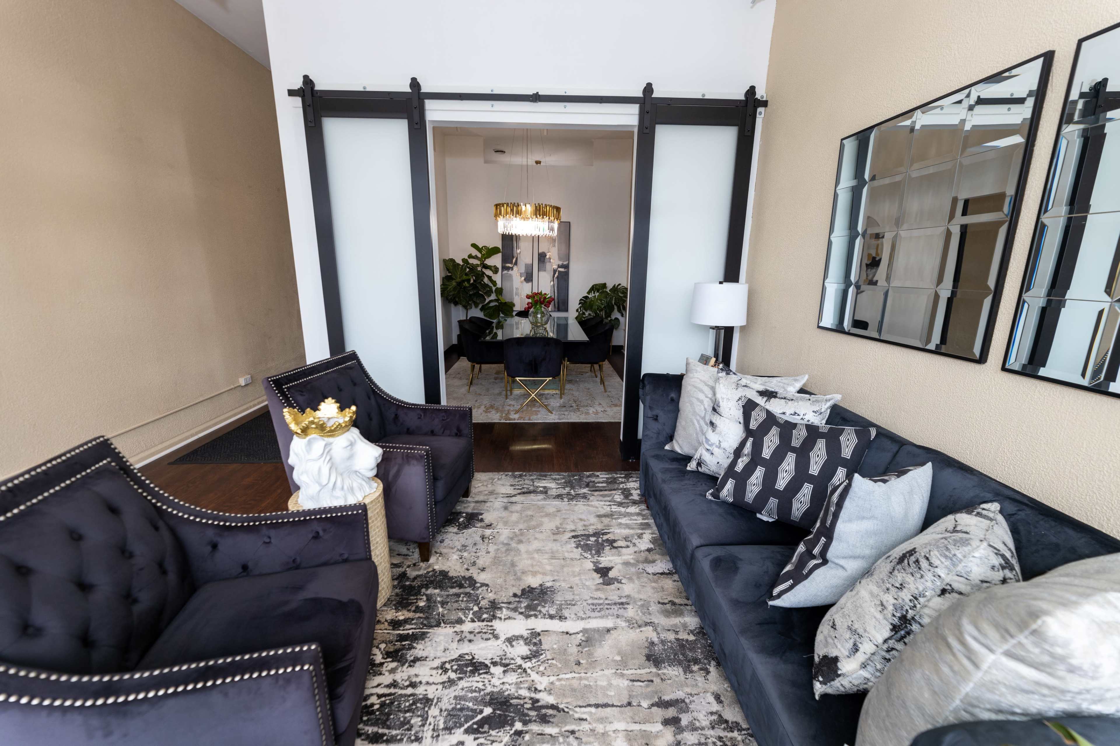 The scene features a stylish living area with two armchairs and a couch, adorned with decorative pillows, leading to a room with a chandelier and plants behind glass sliding doors.