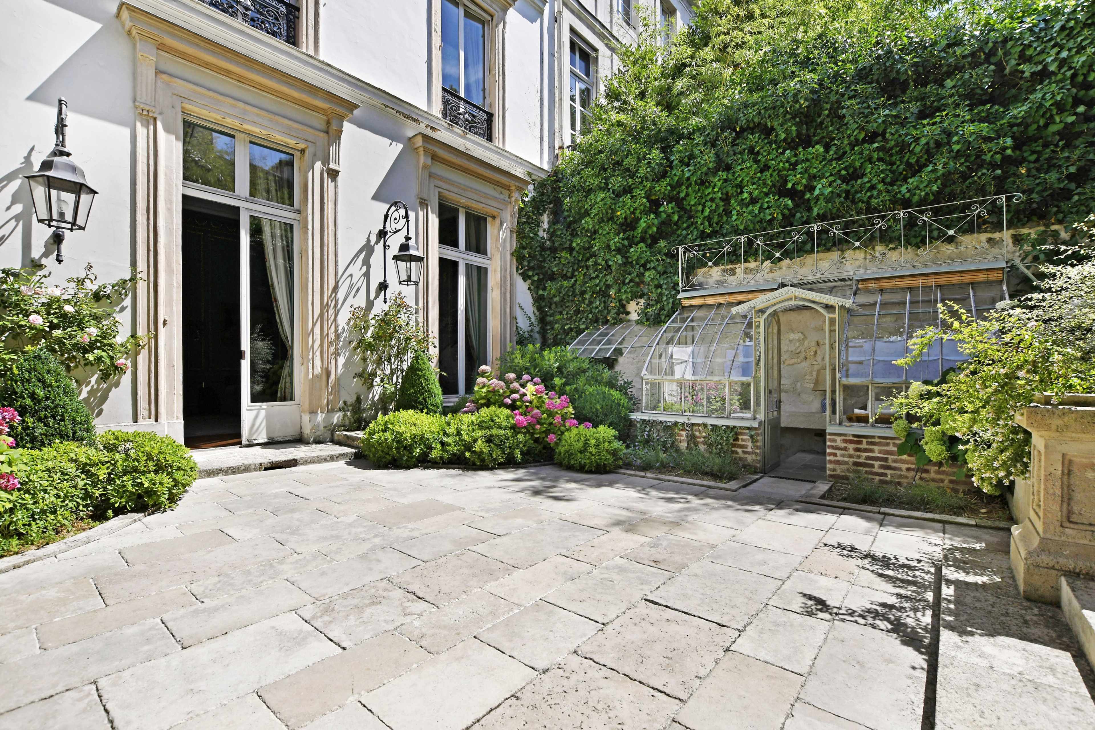A stone-paved courtyard is flanked by greenery and features a glass greenhouse.