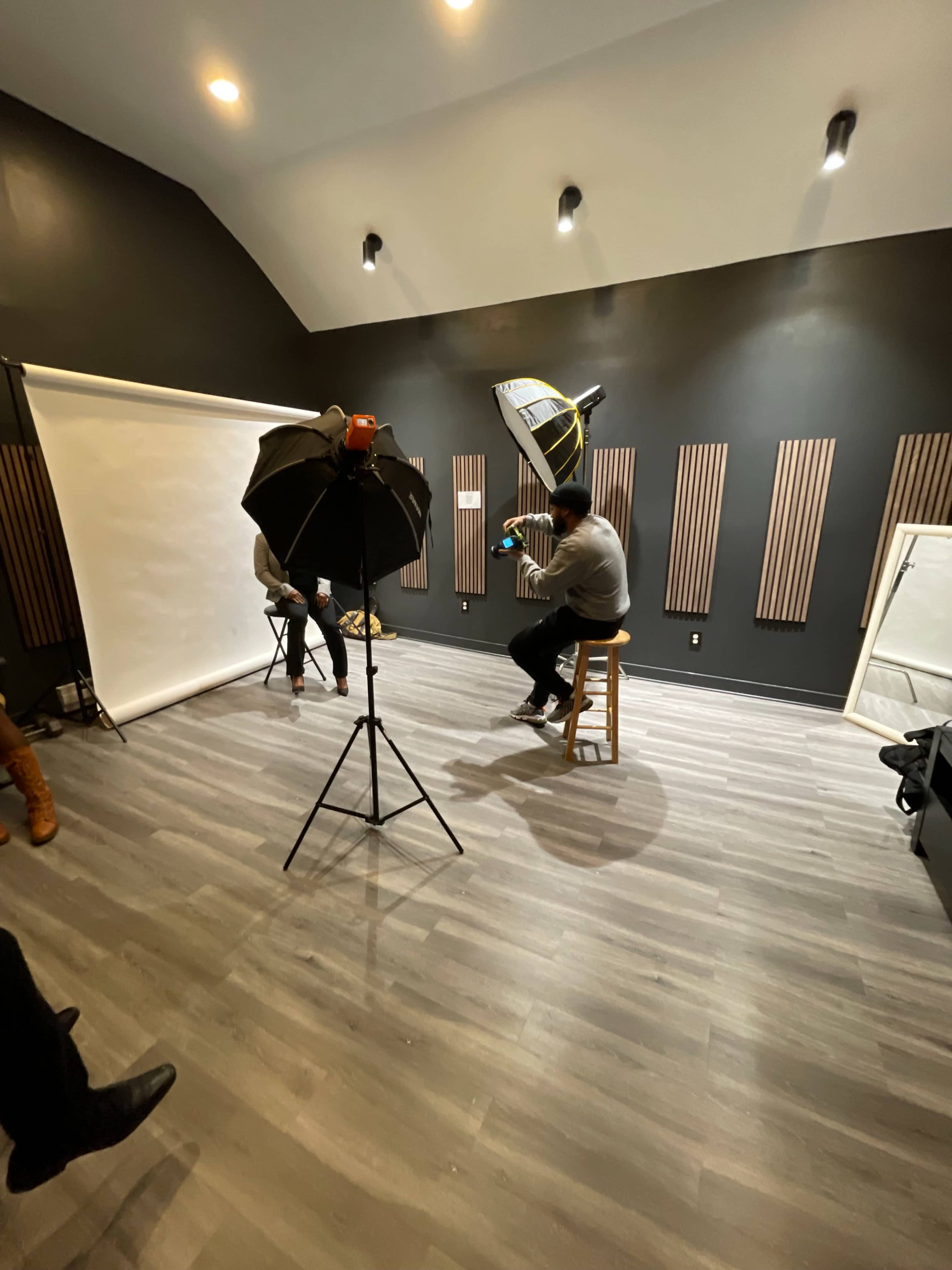 The scene shows a photography studio with a backdrop setup, lighting equipment, and two individuals seated for a photoshoot.