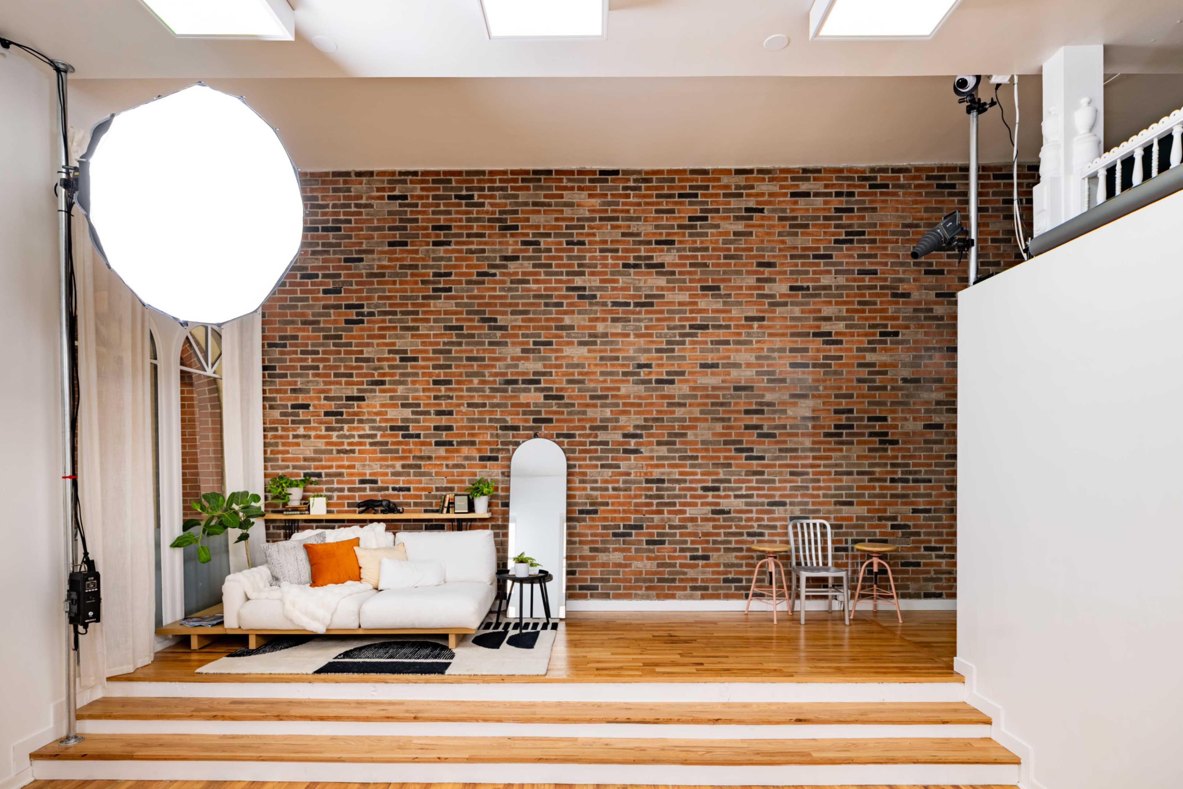 The image shows a modern living room with a brick wall, a white sofa, and wooden steps leading to a small dining area featuring two chairs.