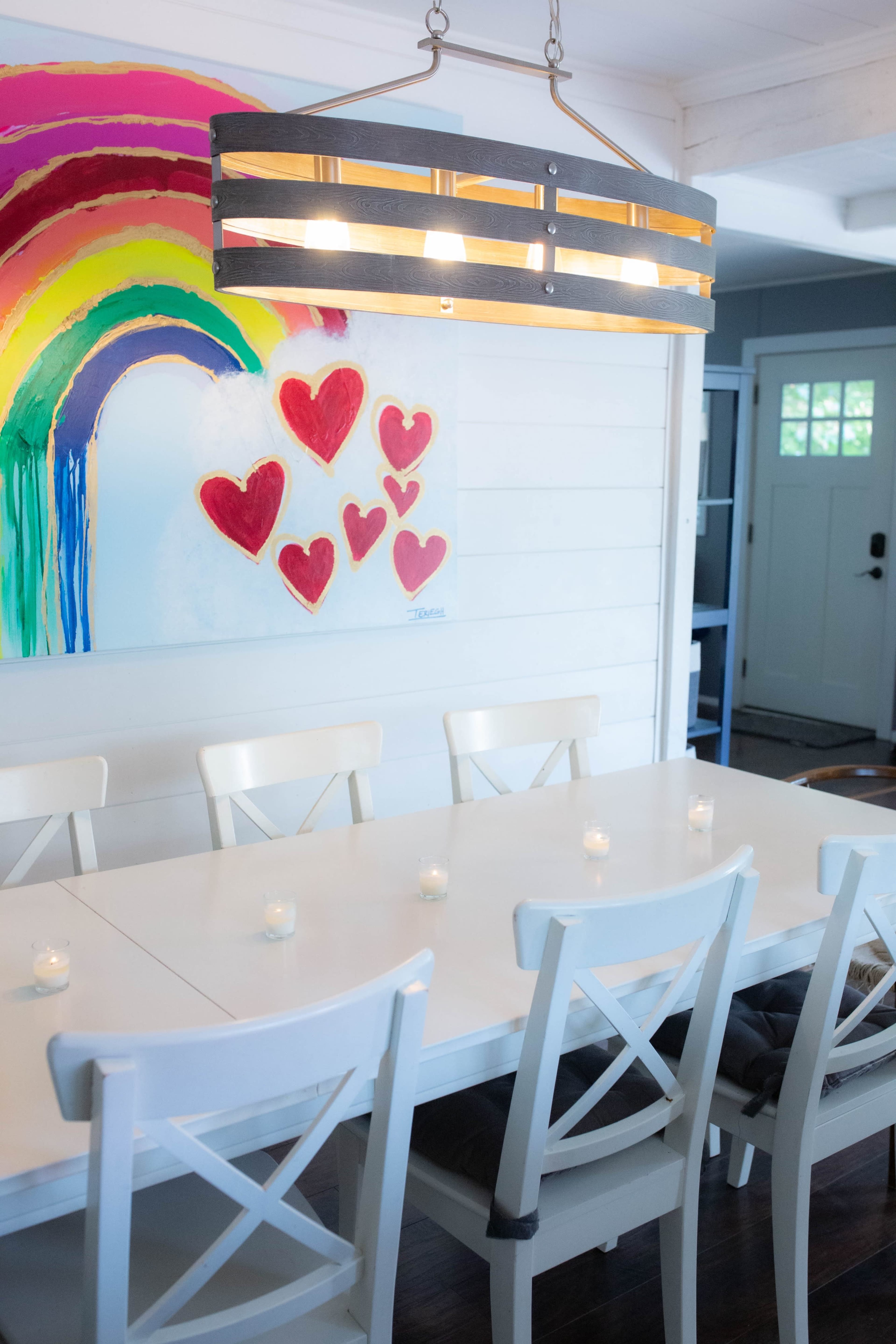 A long white dining table with several chairs is illuminated by a linear light fixture, and a colorful rainbow and heart artwork hangs on the wall.
