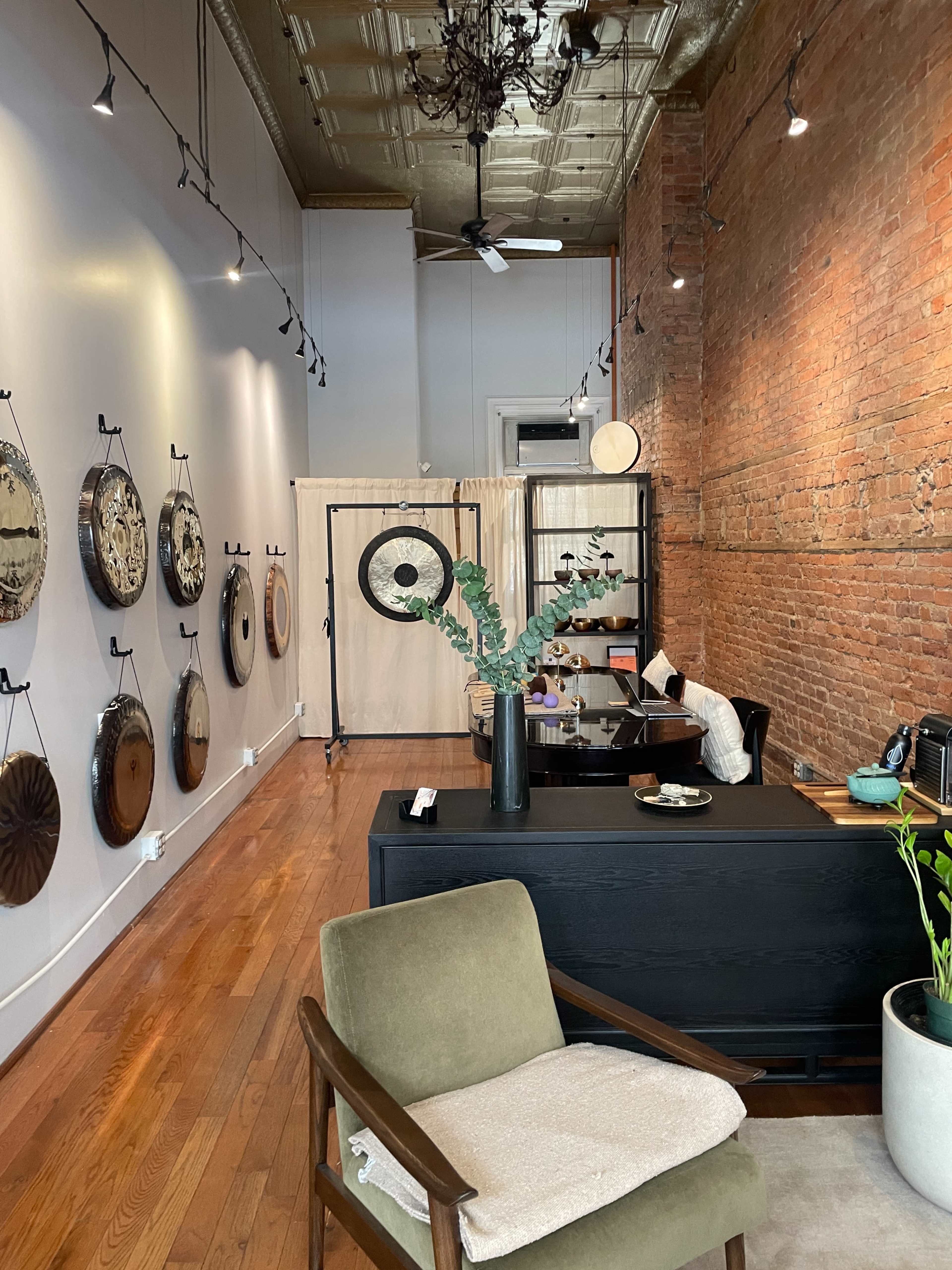 The image shows an art gallery or studio featuring a wooden floor, exposed brick walls, circular art pieces displayed on the walls, and a reception area with a black desk and a green armchair.