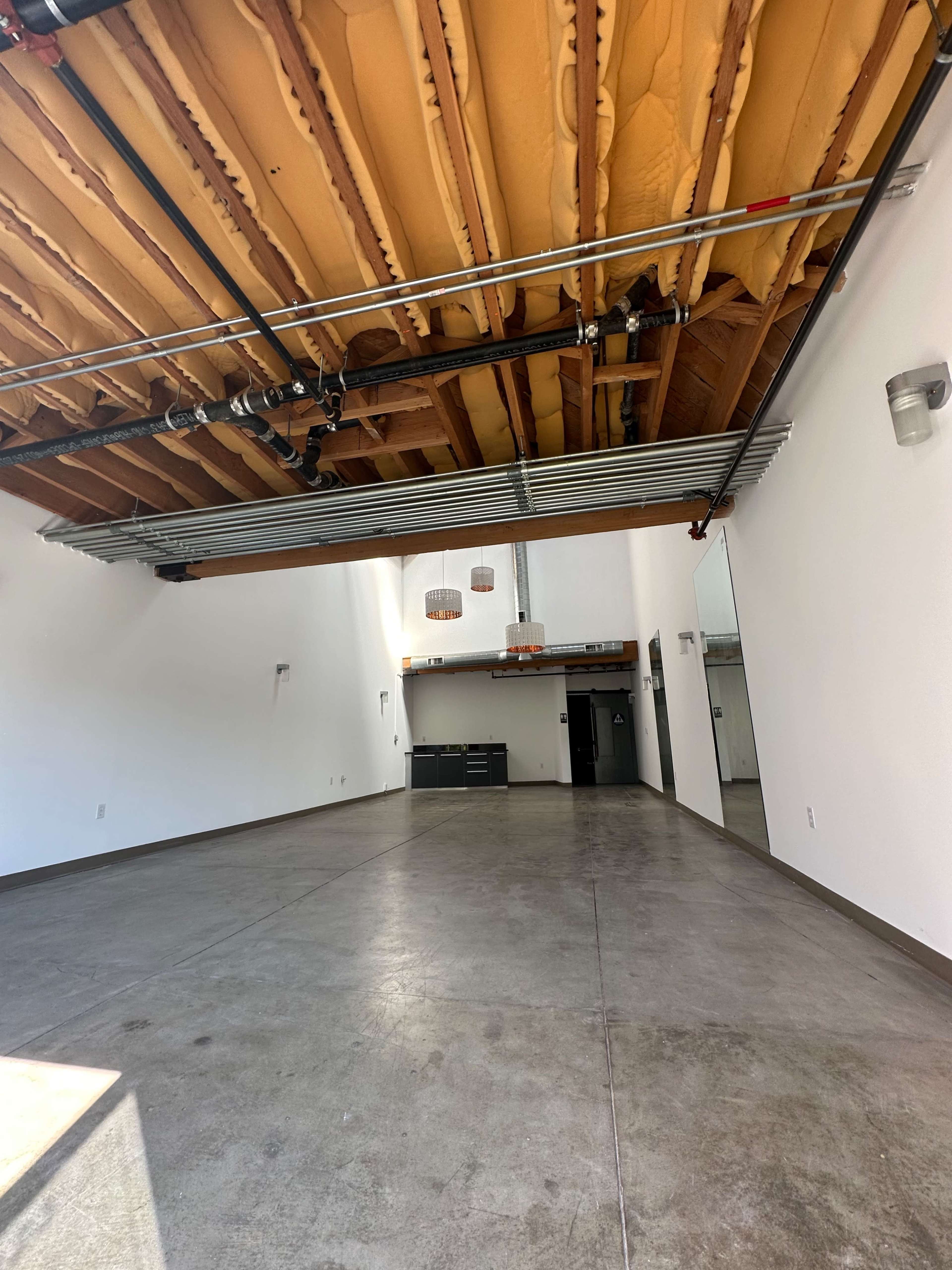 The image shows a spacious interior with exposed wooden beams, polished concrete floors, and minimalist decor, featuring a kitchen area at the back.