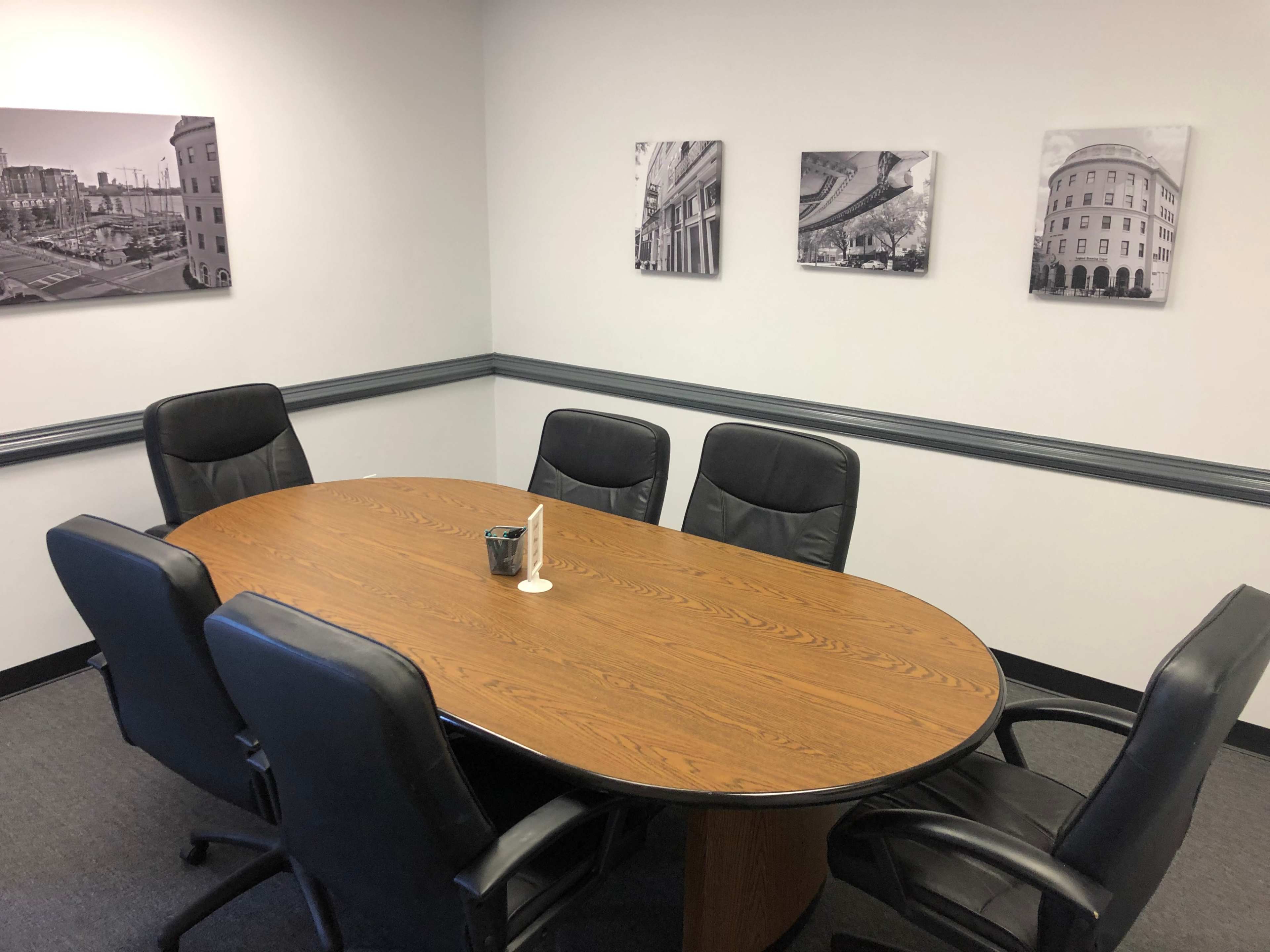 A conference room features a large oval wooden table surrounded by six black office chairs, with black-and-white photographs hanging on the wall.