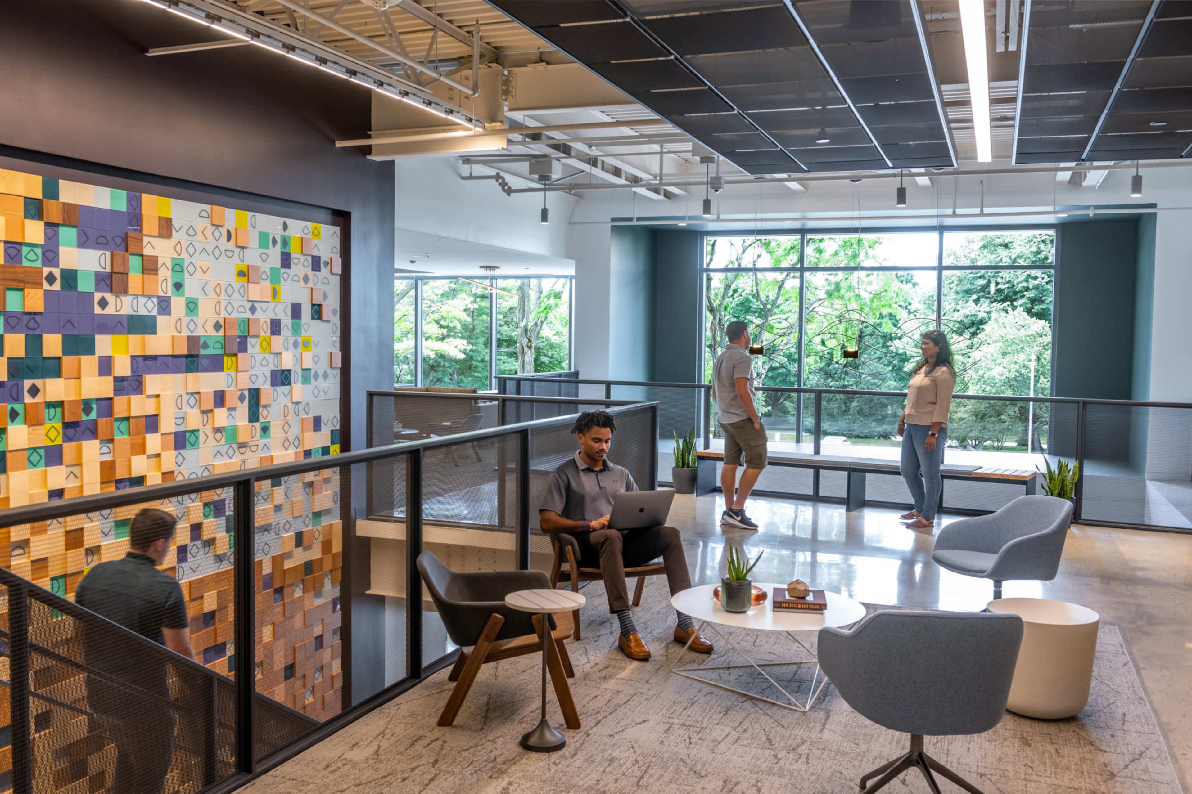 A modern office space with a colorful wall mural, where a person works on a laptop while others engage in conversation and stand near large windows overlooking greenery.