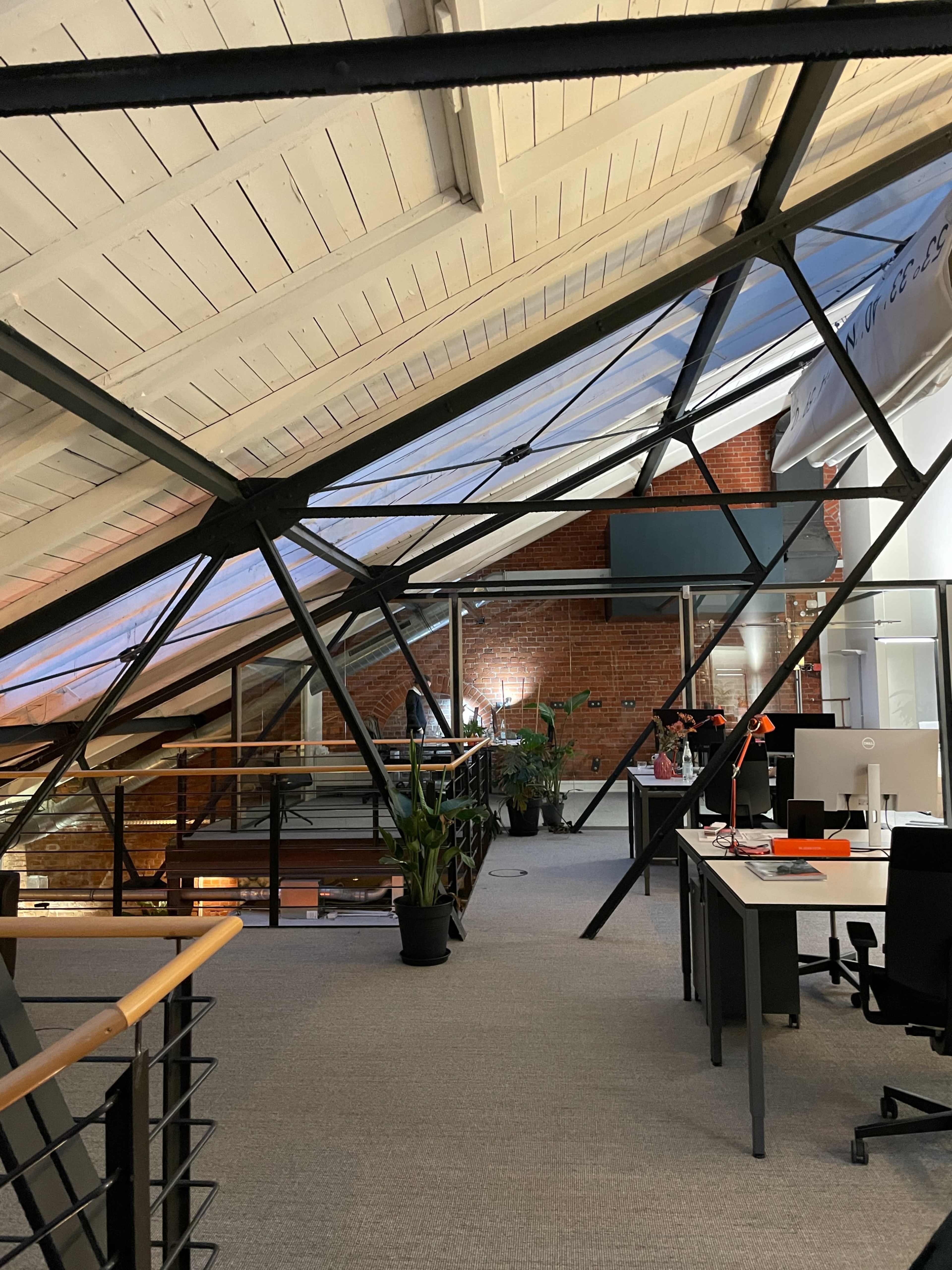 The image shows a modern office space with a lofted ceiling, large windows, metal beams, and workstations arranged around a central area with greenery.