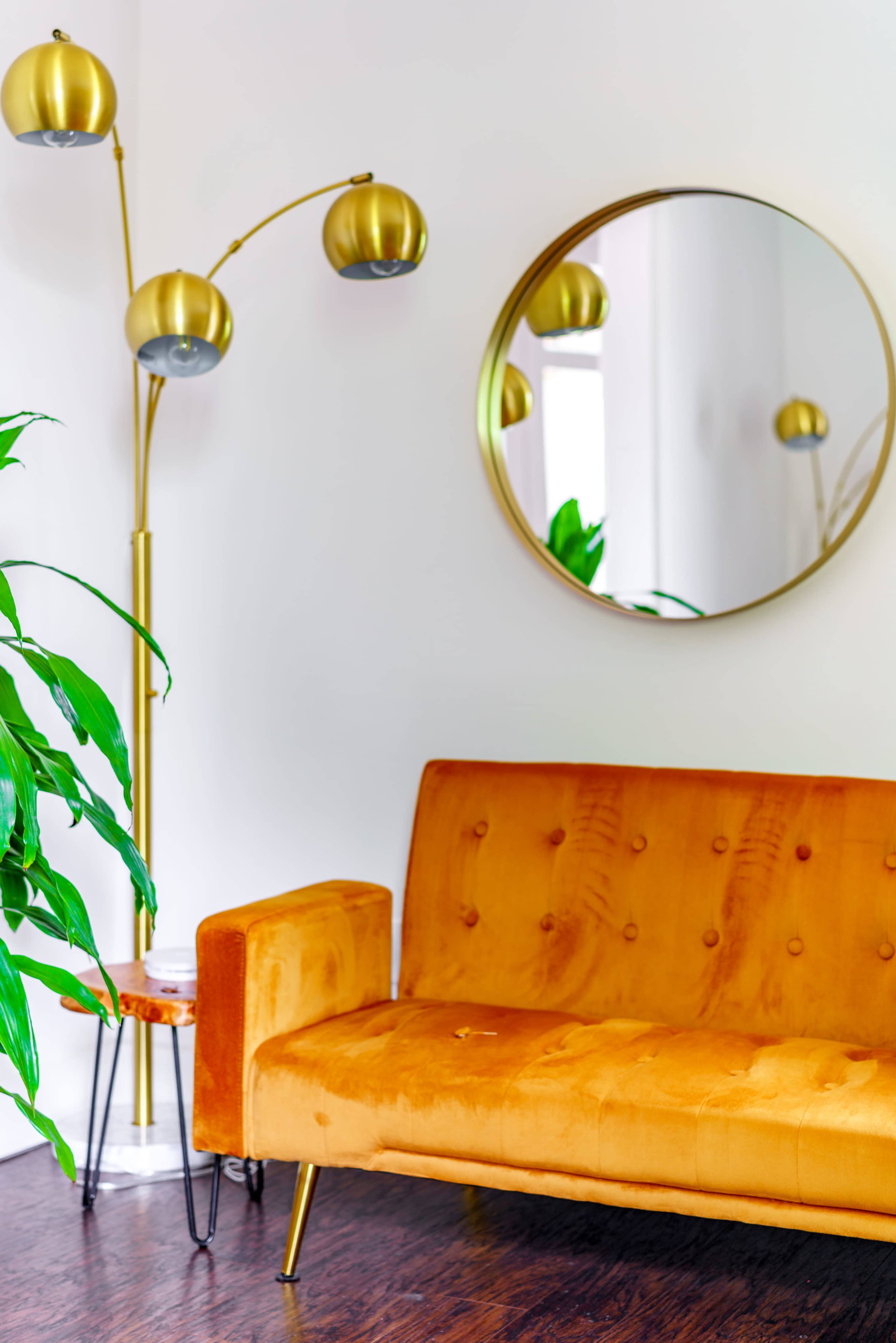 The image shows a modern living room featuring an orange velvet sofa, a gold arc floor lamp, a round mirror on the wall, and a green plant in the corner.