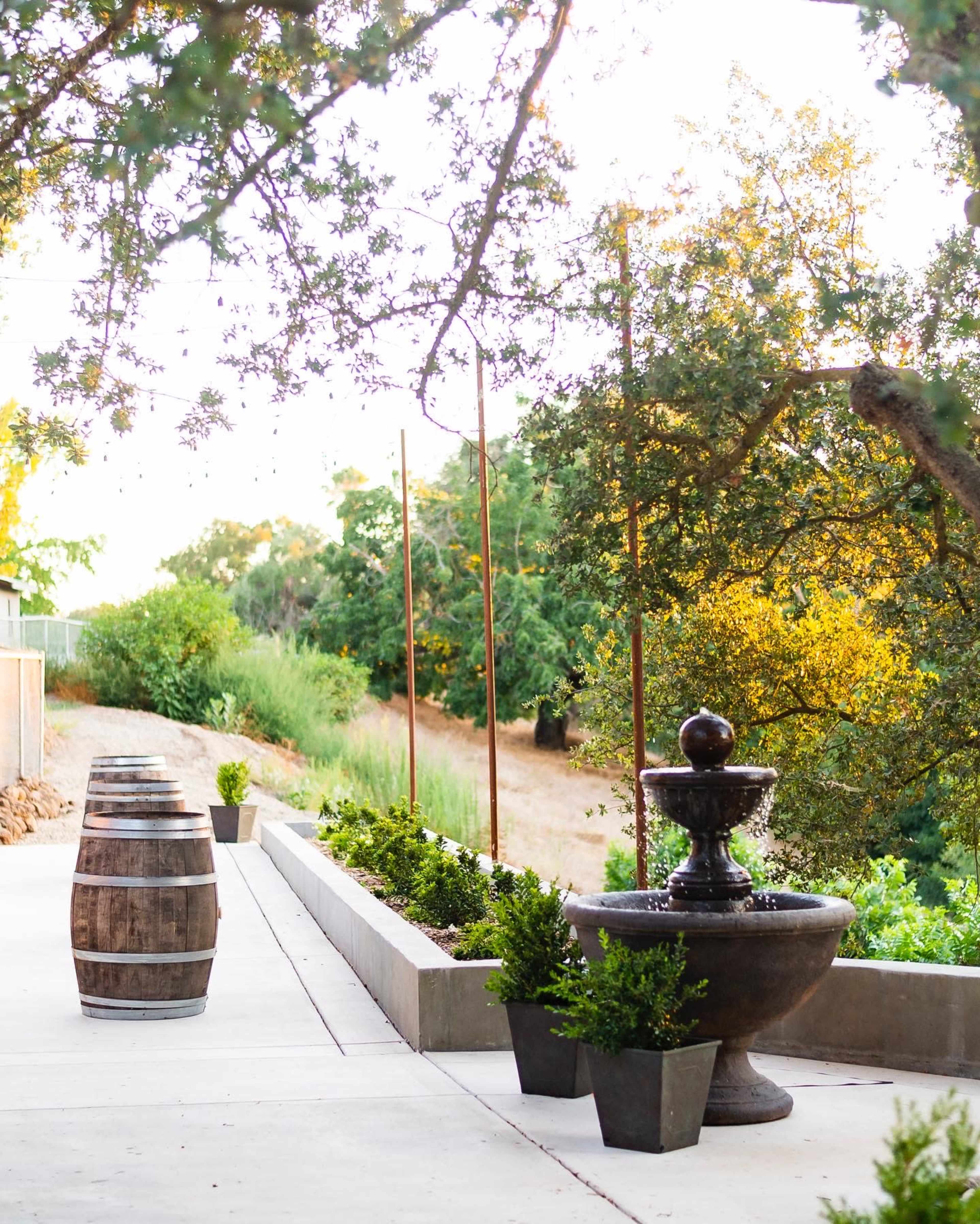 A pathway lined with planters and a stone fountain, flanked by wooden barrels and surrounded by trees.
