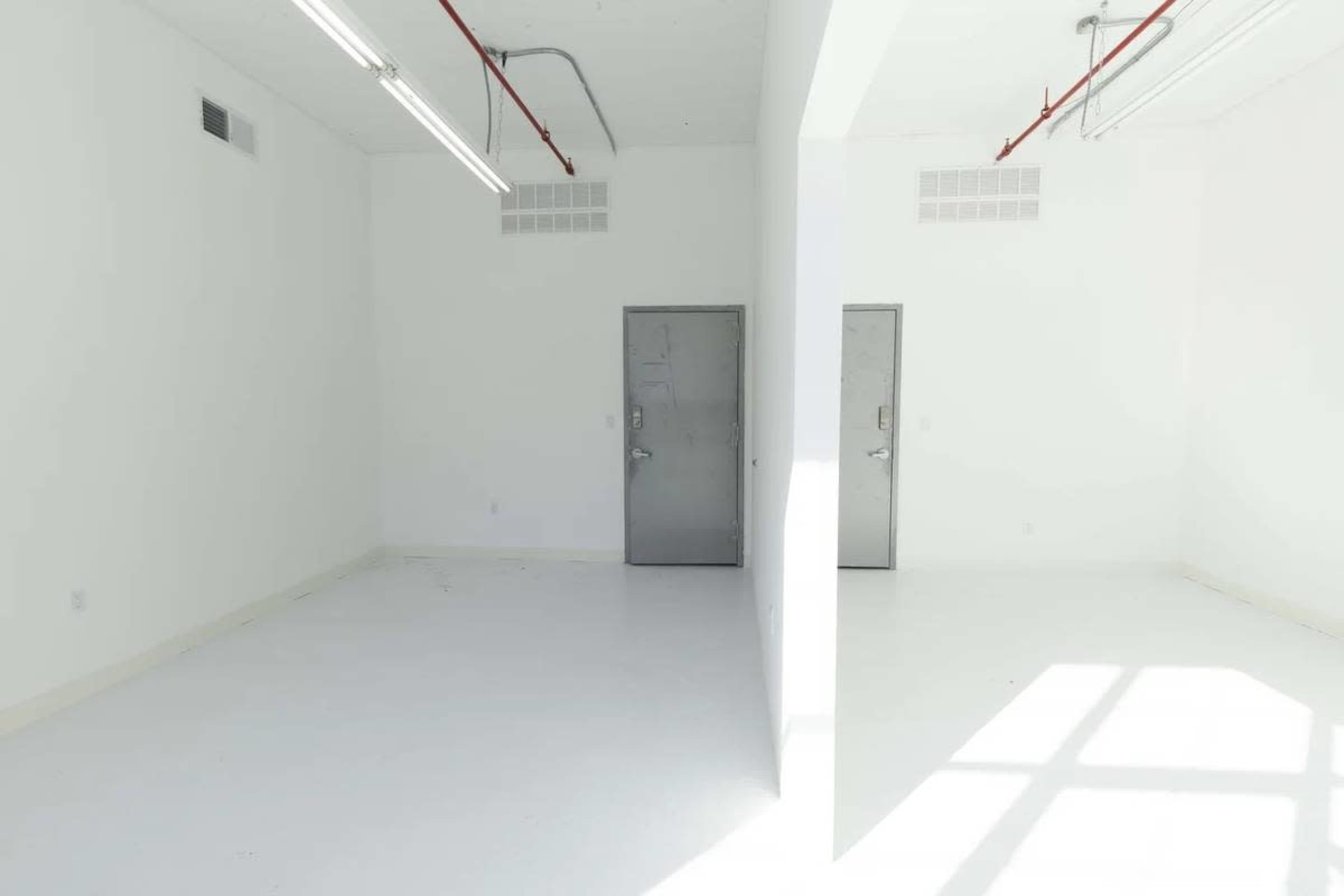 The image shows a large, empty room divided by a wall, featuring two gray doors and bright, artificial lighting.