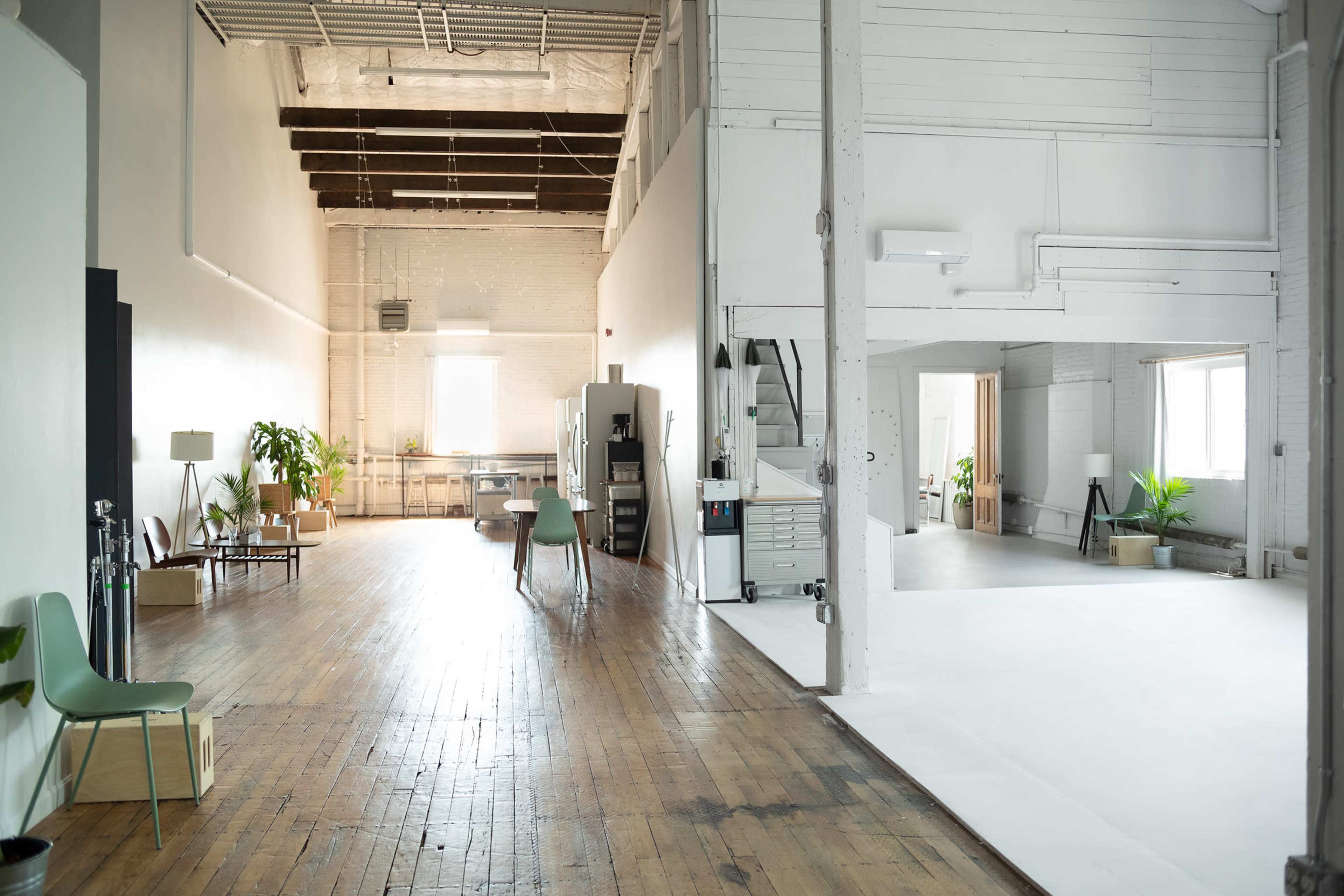 The image shows a spacious, modern studio with hardwood floors, bright white walls, and various plants, divided into two areas: one with workstations and furniture, and the other an open, blank space.