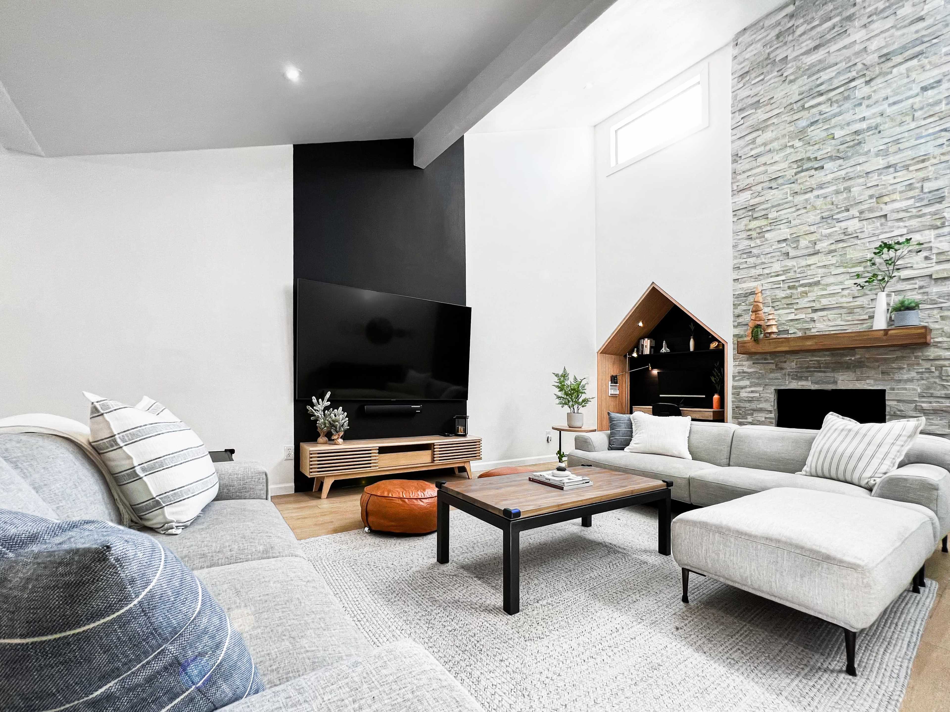 A modern living room features a large TV mounted on a black wall, a stone fireplace, and a mix of light-colored sofas arranged around a coffee table.