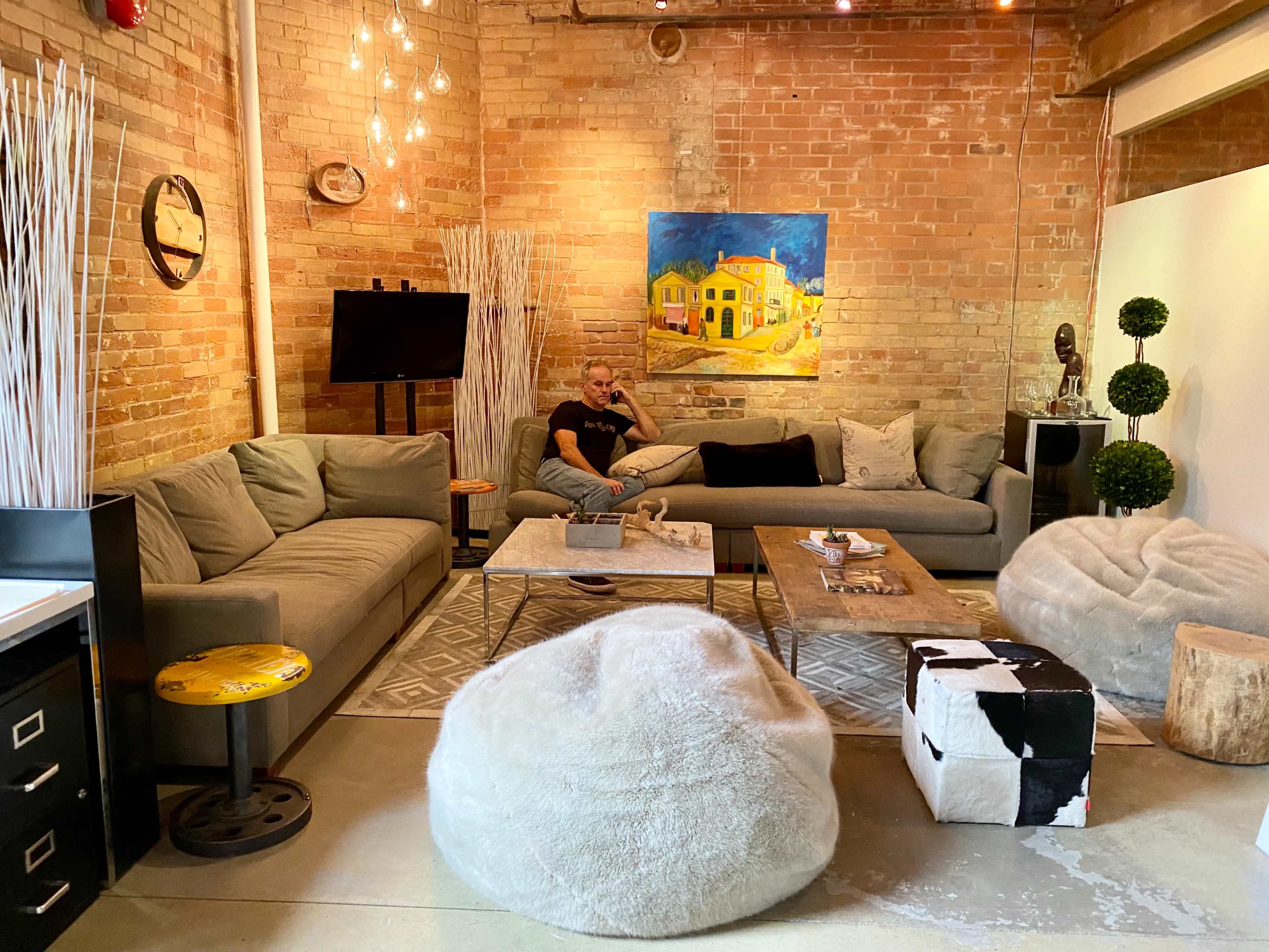 A modern living room features a gray sectional sofa, a wooden coffee table, and bean bag chairs, with brick walls and artwork enhancing the decor.